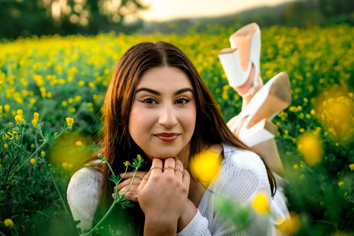 Young woman lying in a vibrant yellow wildflower field, smiling warmly at camera during golden hour.