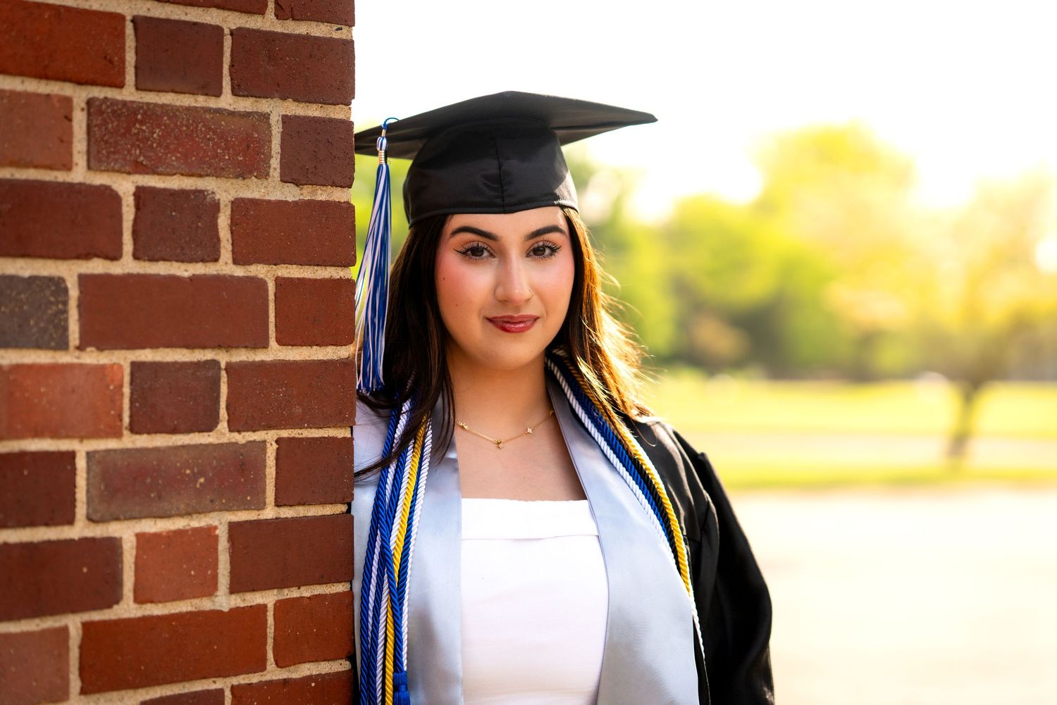 Young woman in black graduation cap and gown with blue and yellow tassel smiling outdoors near brick wall.