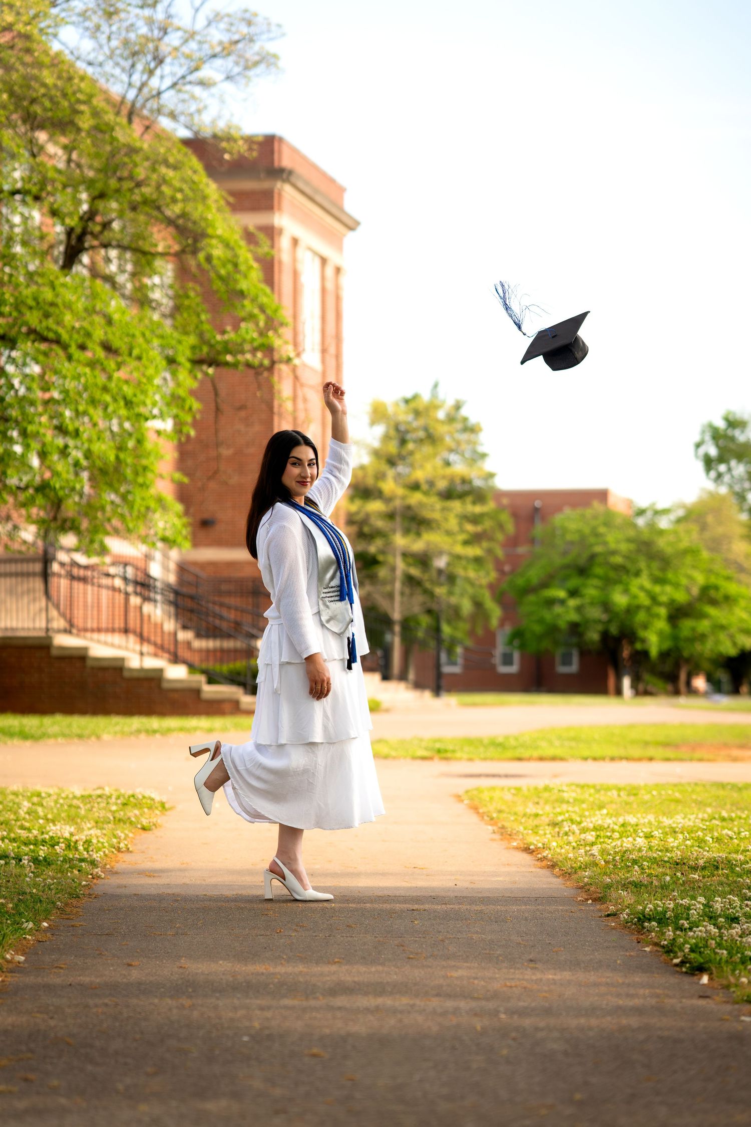 A joyful graduate in white gown tosses her cap in the air on a tree-lined campus walkway.