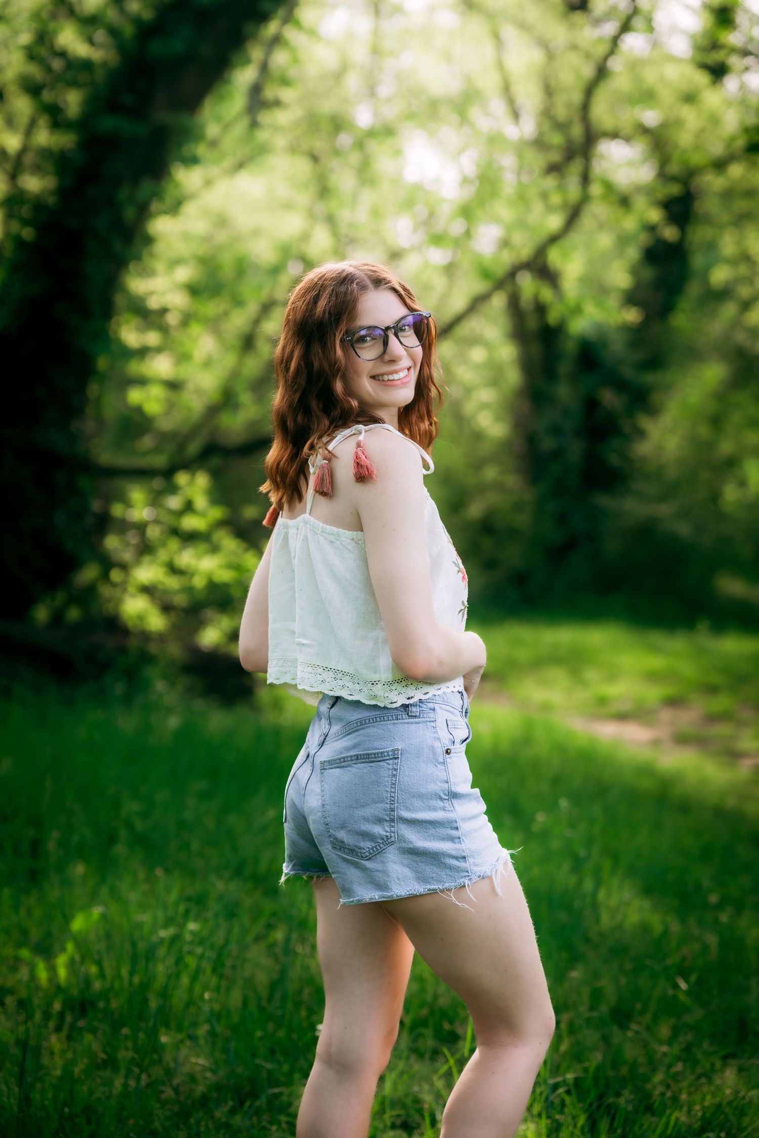Smiling redhead woman in glasses and denim shorts posing in a lush green forest path.