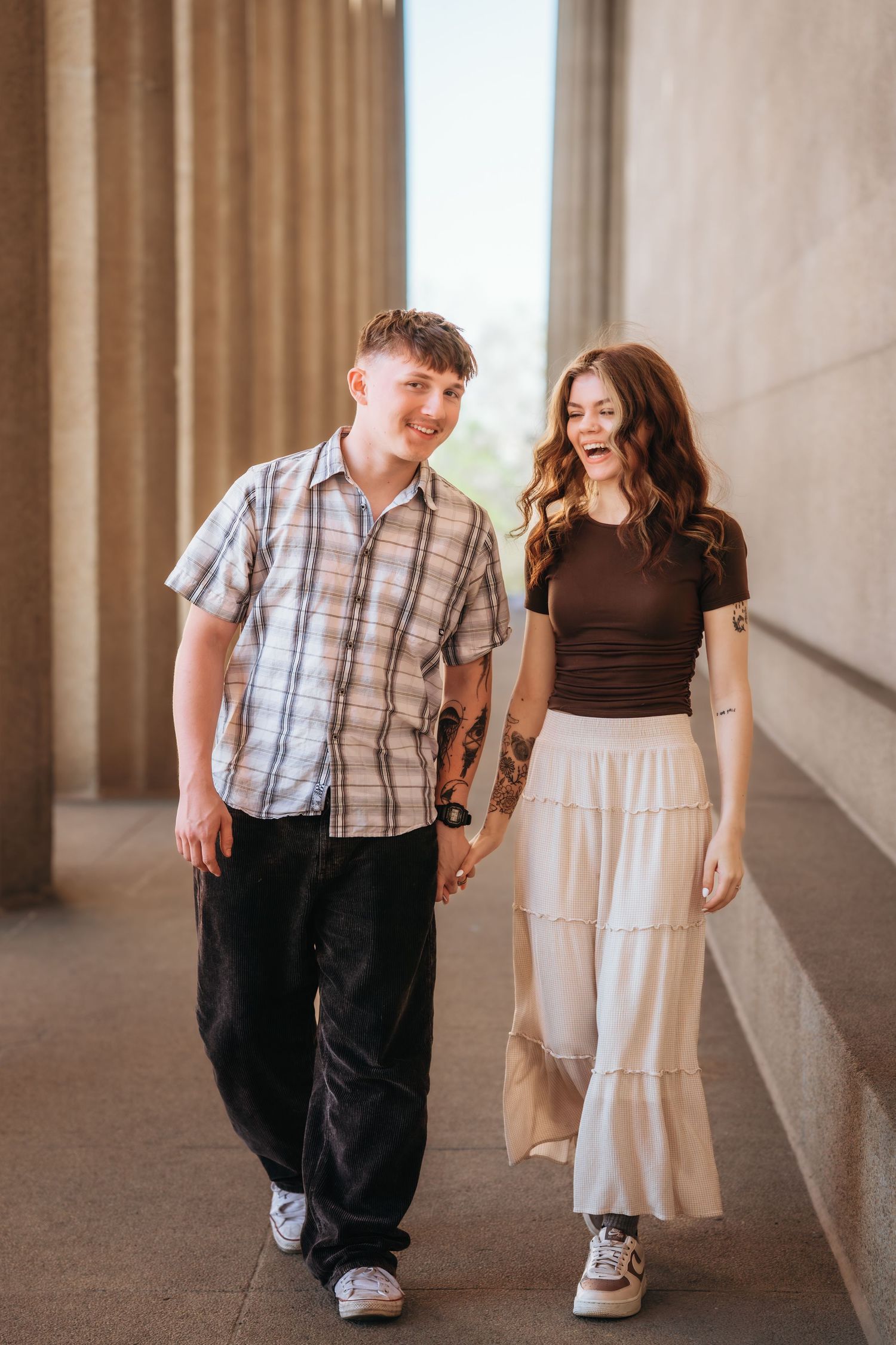 A young couple holds hands and laughs while walking between stone columns, dressed in casual summer outfits.