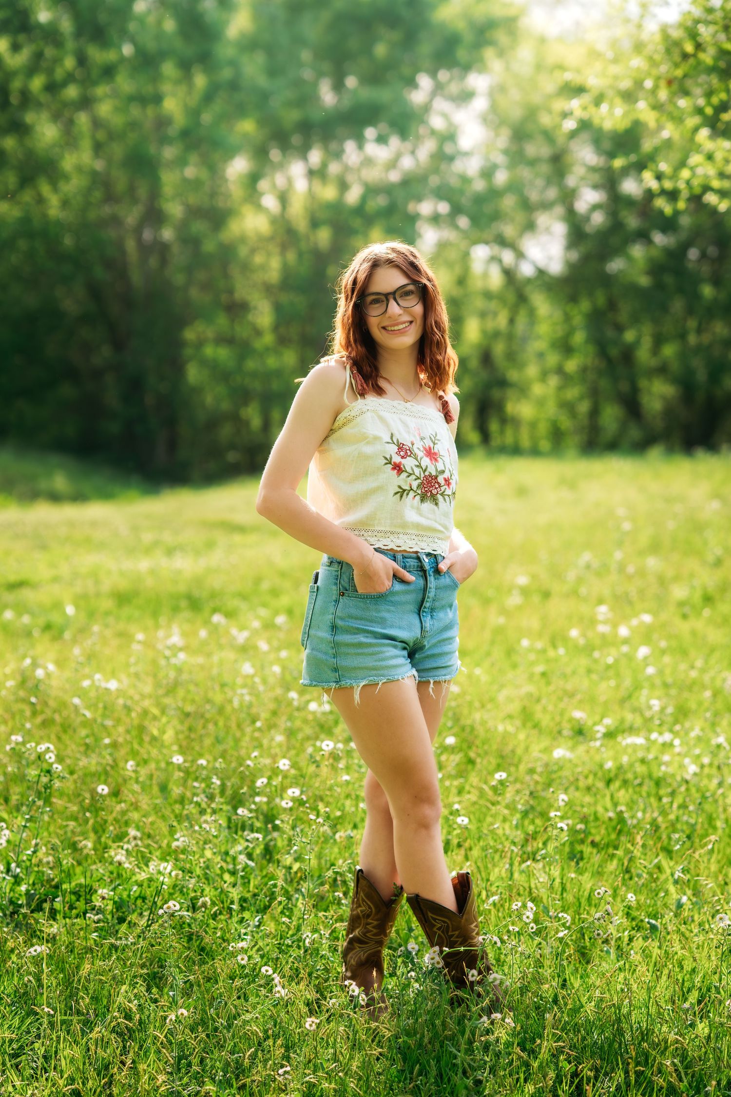 Young woman in floral top, denim shorts, and cowboy boots standing in a sunny green meadow with wildflowers.