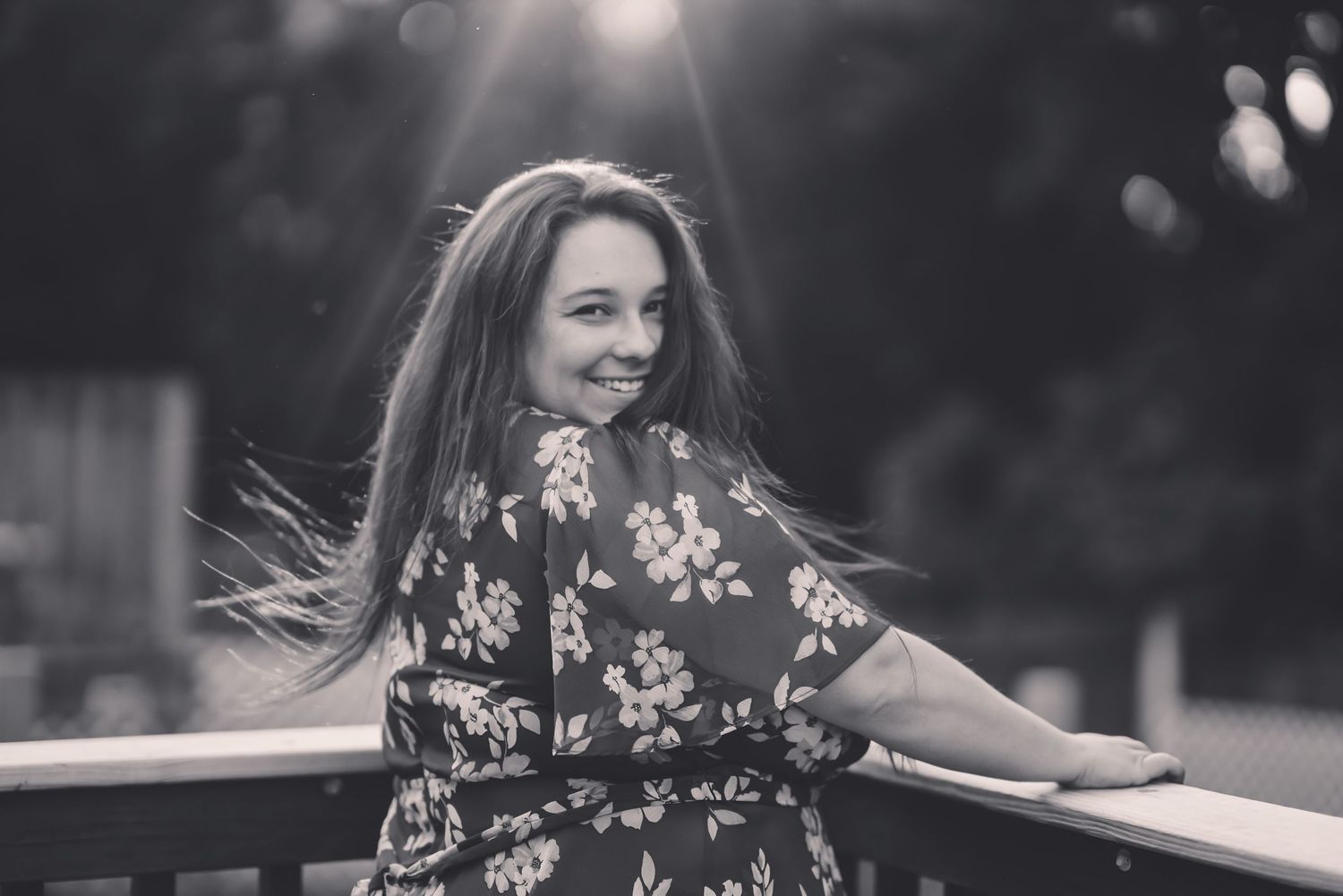 Young woman in floral dress smiling over shoulder, black and white portrait with sunlight bokeh background.