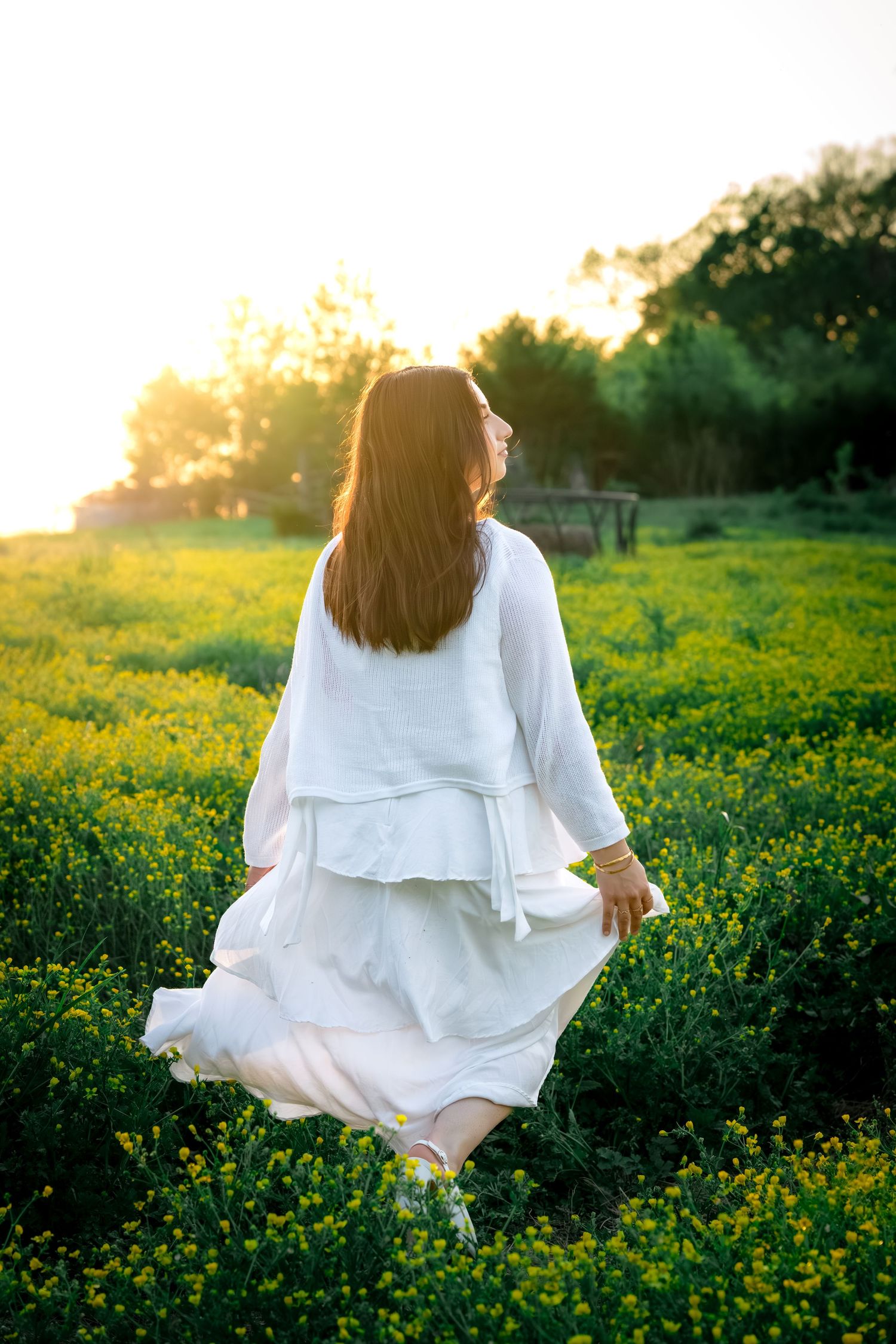 Woman in white tiered dress twirling in yellow flower field bathed in golden sunset light.