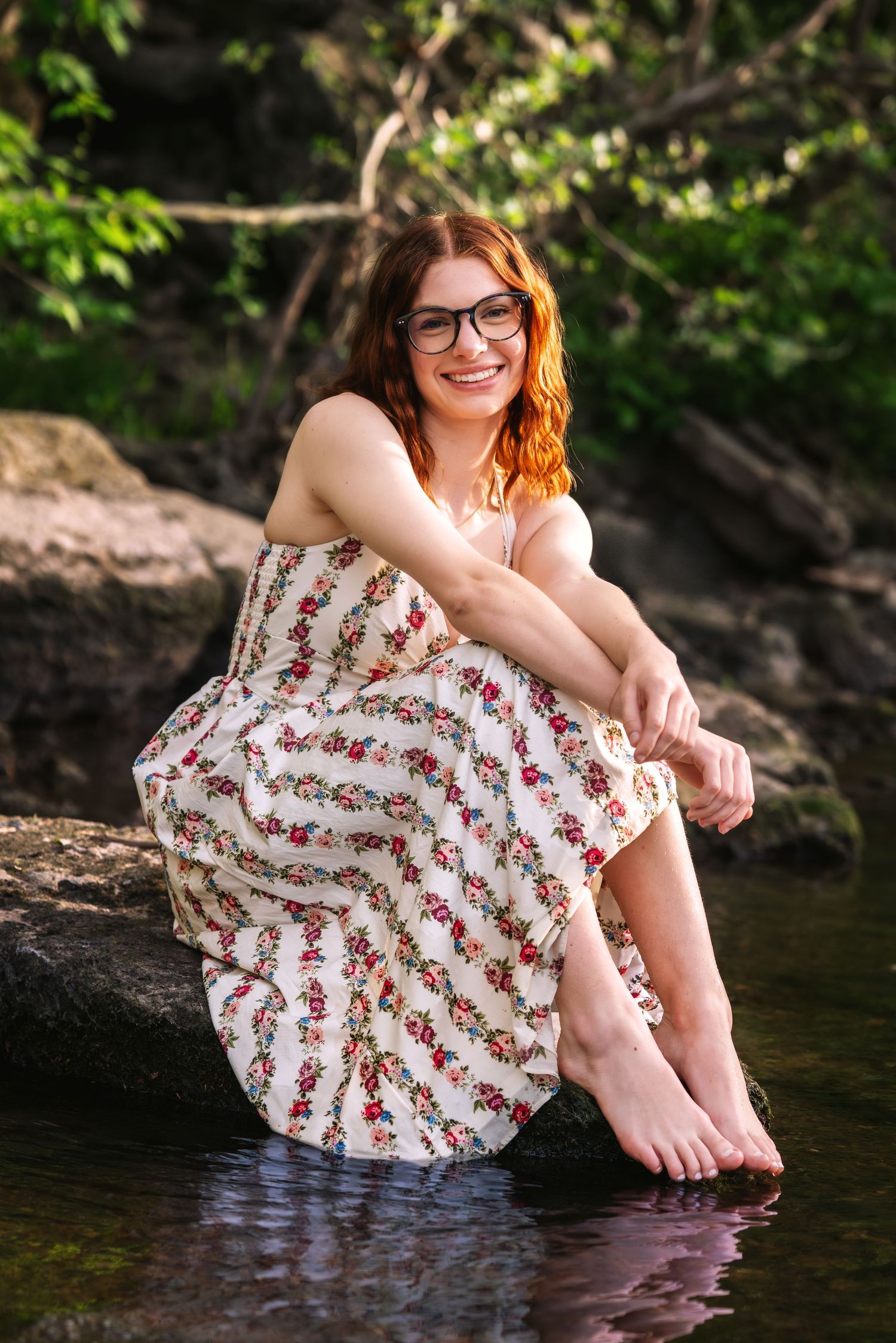 Smiling redhead woman in floral dress sitting barefoot on rocks by a peaceful stream outdoors.