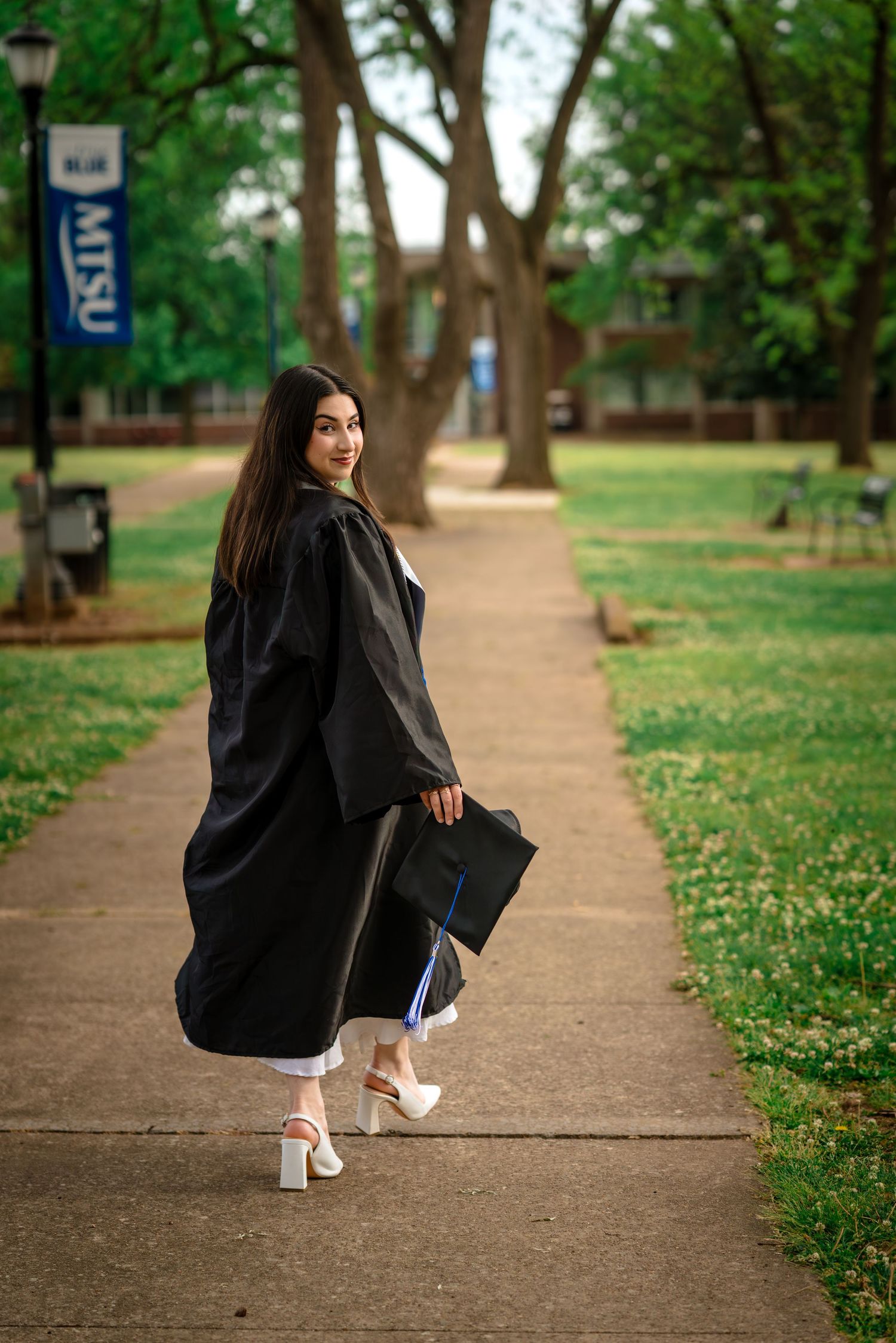 A young woman in black graduation gown and cap walks on a campus path at MTSU, smiling over her shoulder.