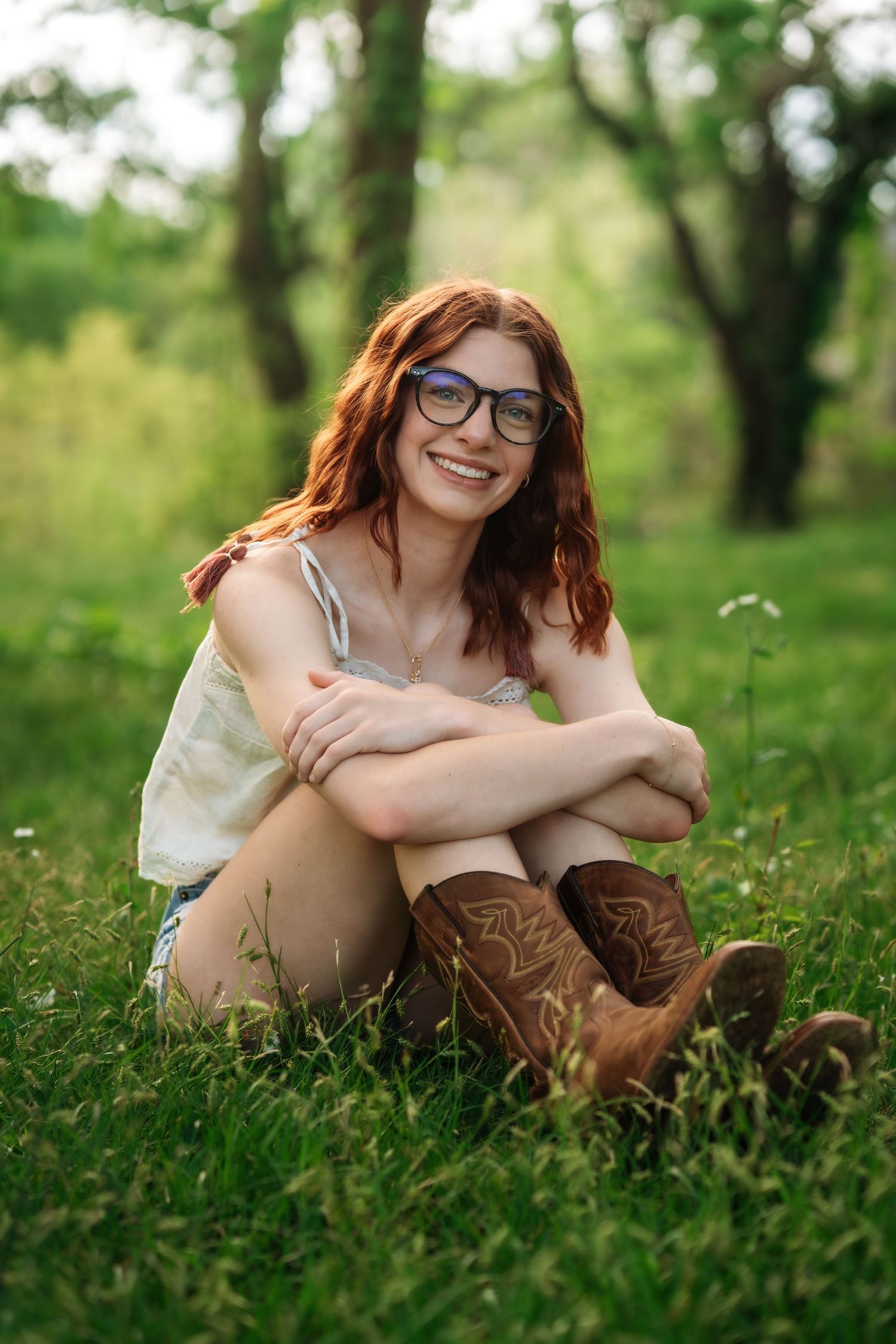 Cheerful young redhead woman in glasses and brown cowboy boots sitting cross-legged on lush green grass outdoors.
