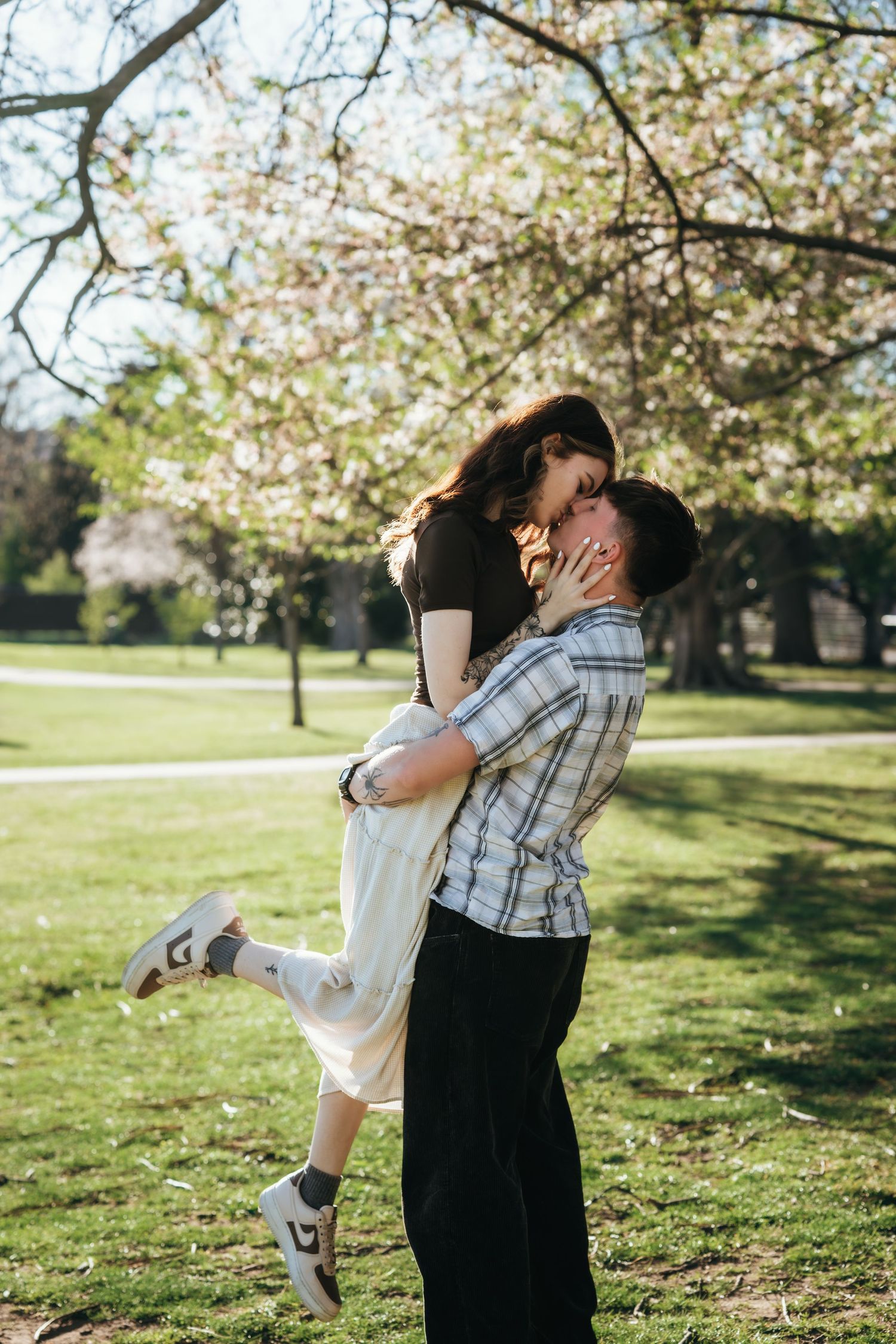 Couple sharing a romantic lift and kiss under blooming cherry tree in a sunny green park.