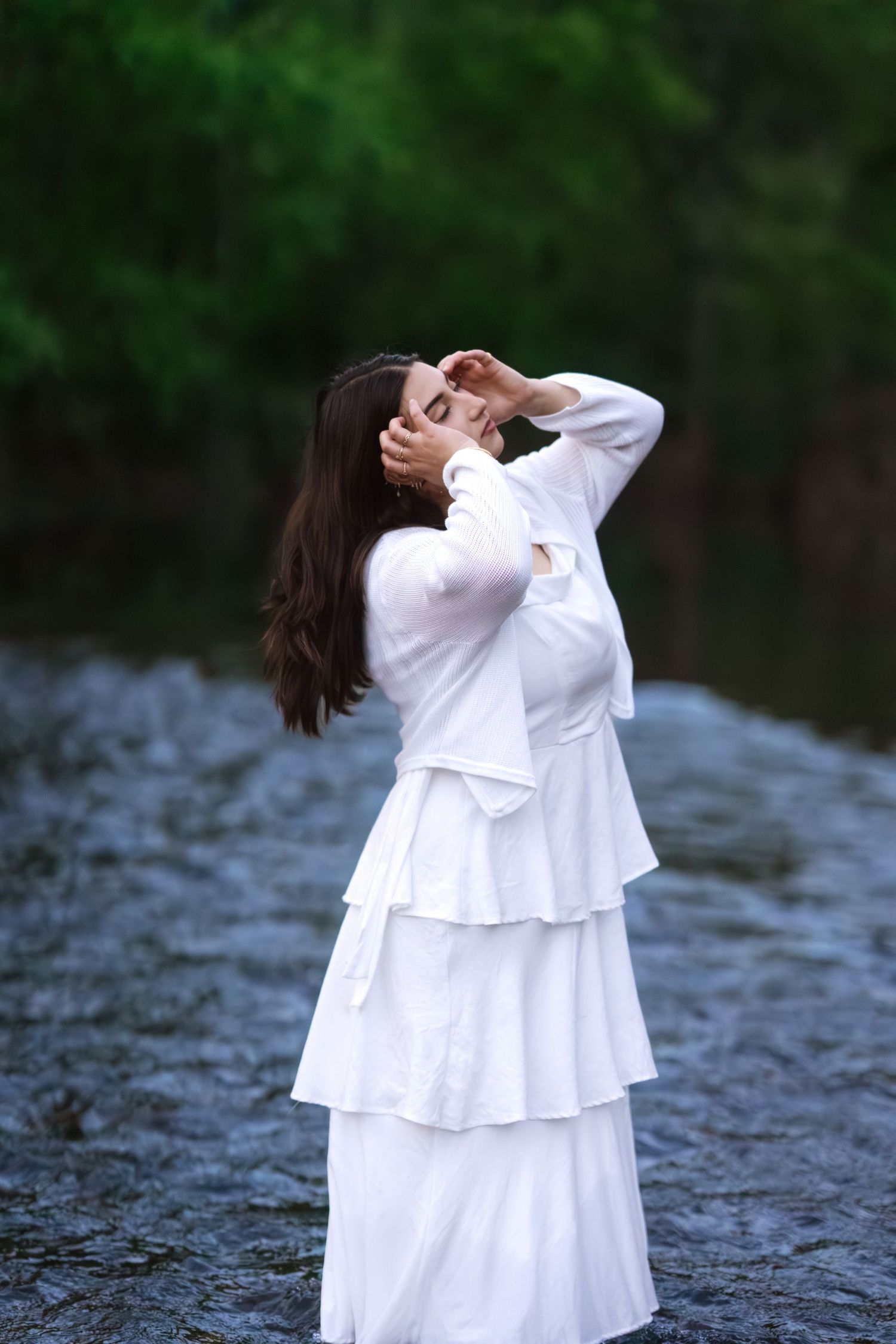 Young woman in white layered dress poses gracefully by rocky stream in lush green forest.