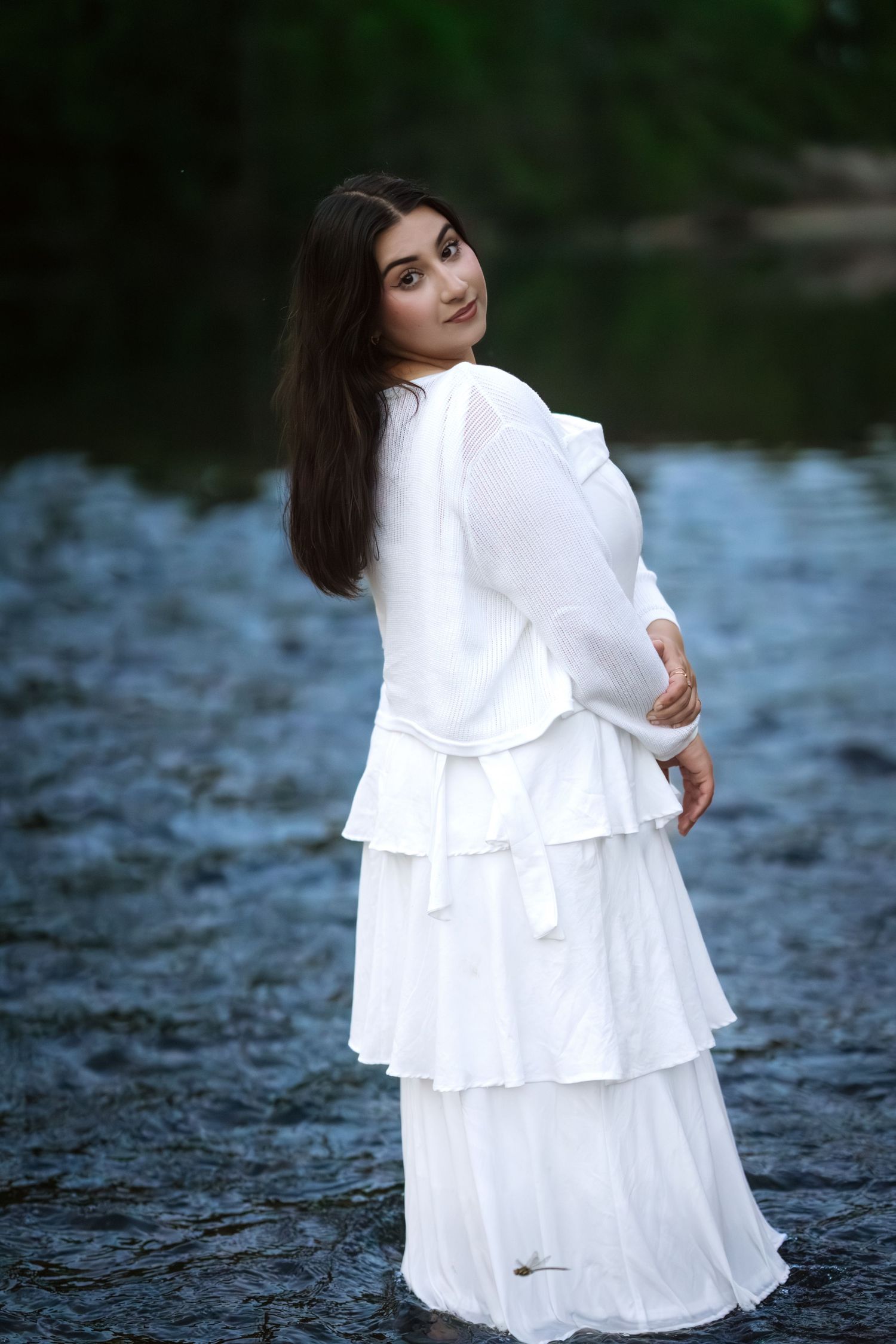 Elegant young woman in white ruffled tiered dress stands in river turning to smile over shoulder.