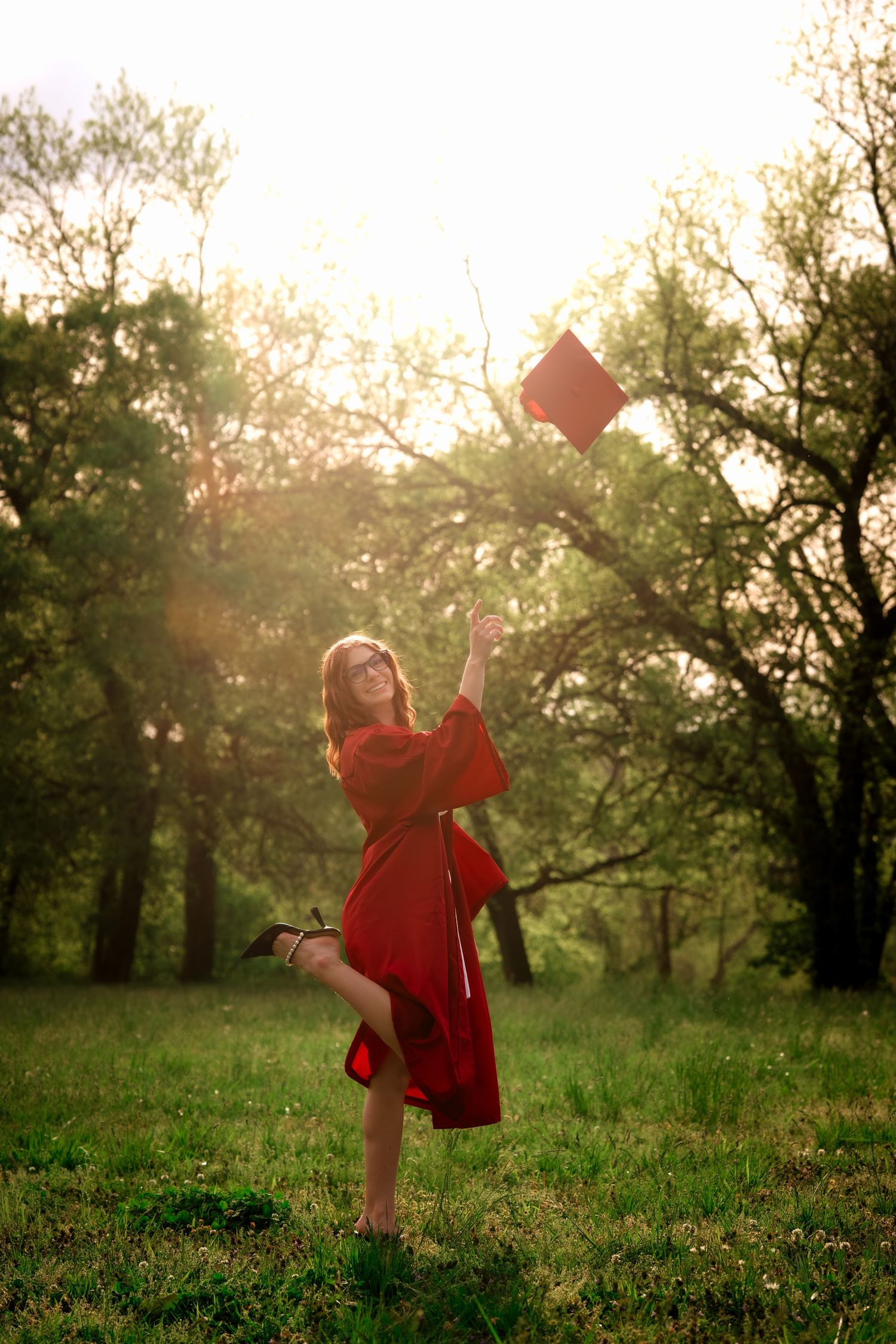 Joyful graduate in red gown tossing her cap in a sunlit outdoor setting.
