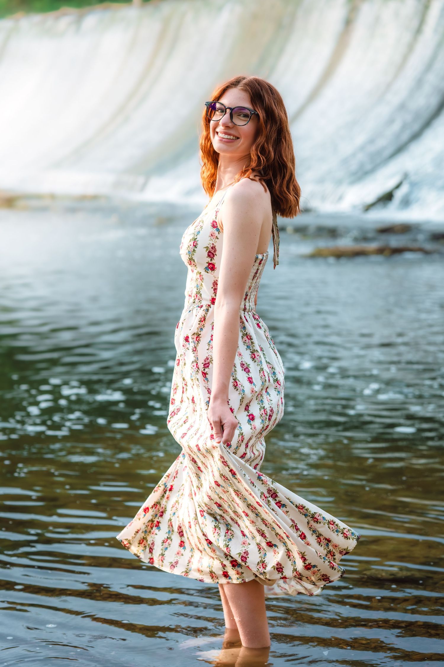 Redhead woman in floral maxi dress standing barefoot in a river near a waterfall.