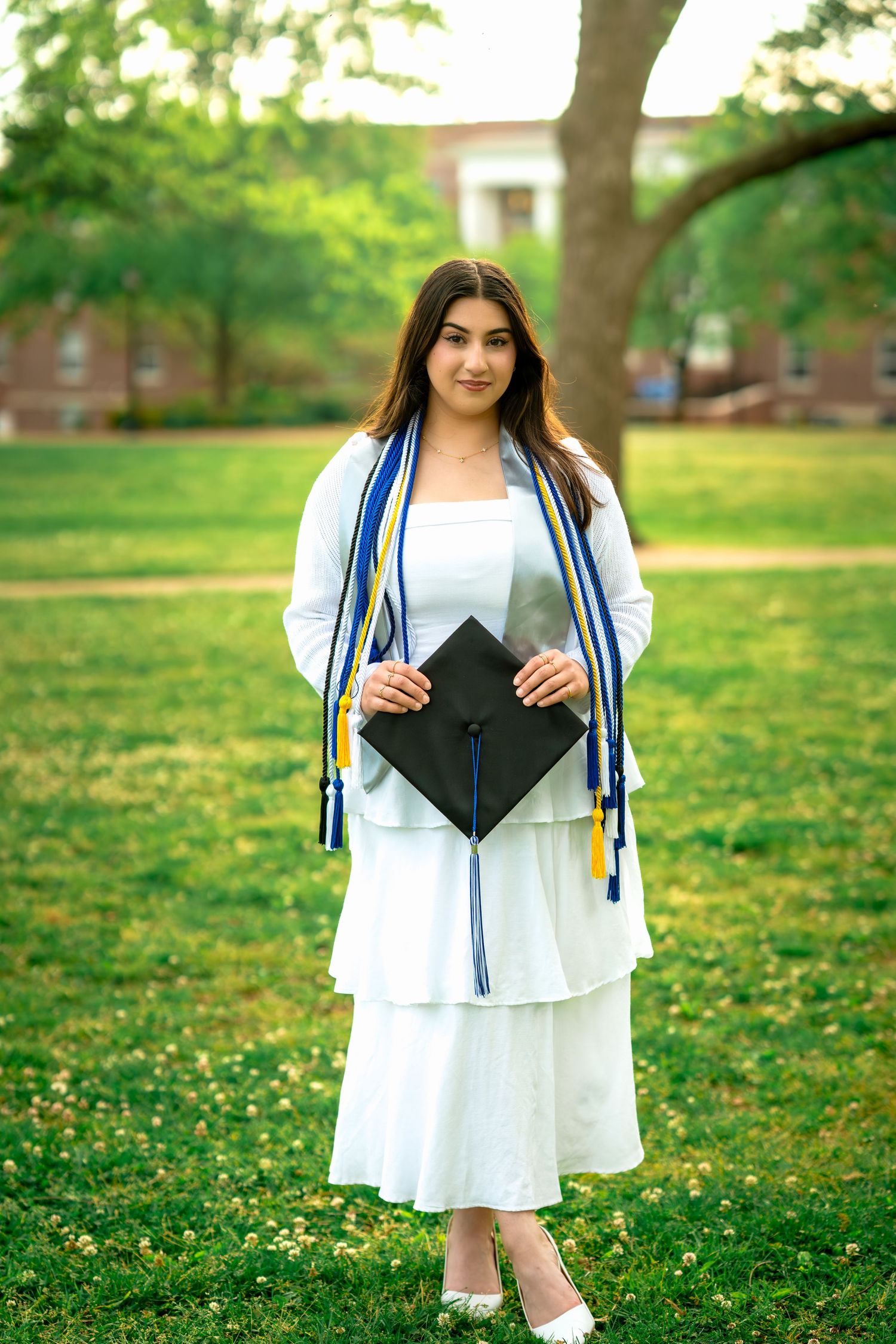 Smiling graduate in white dress holds black cap with blue and gold honor cords on green campus lawn.