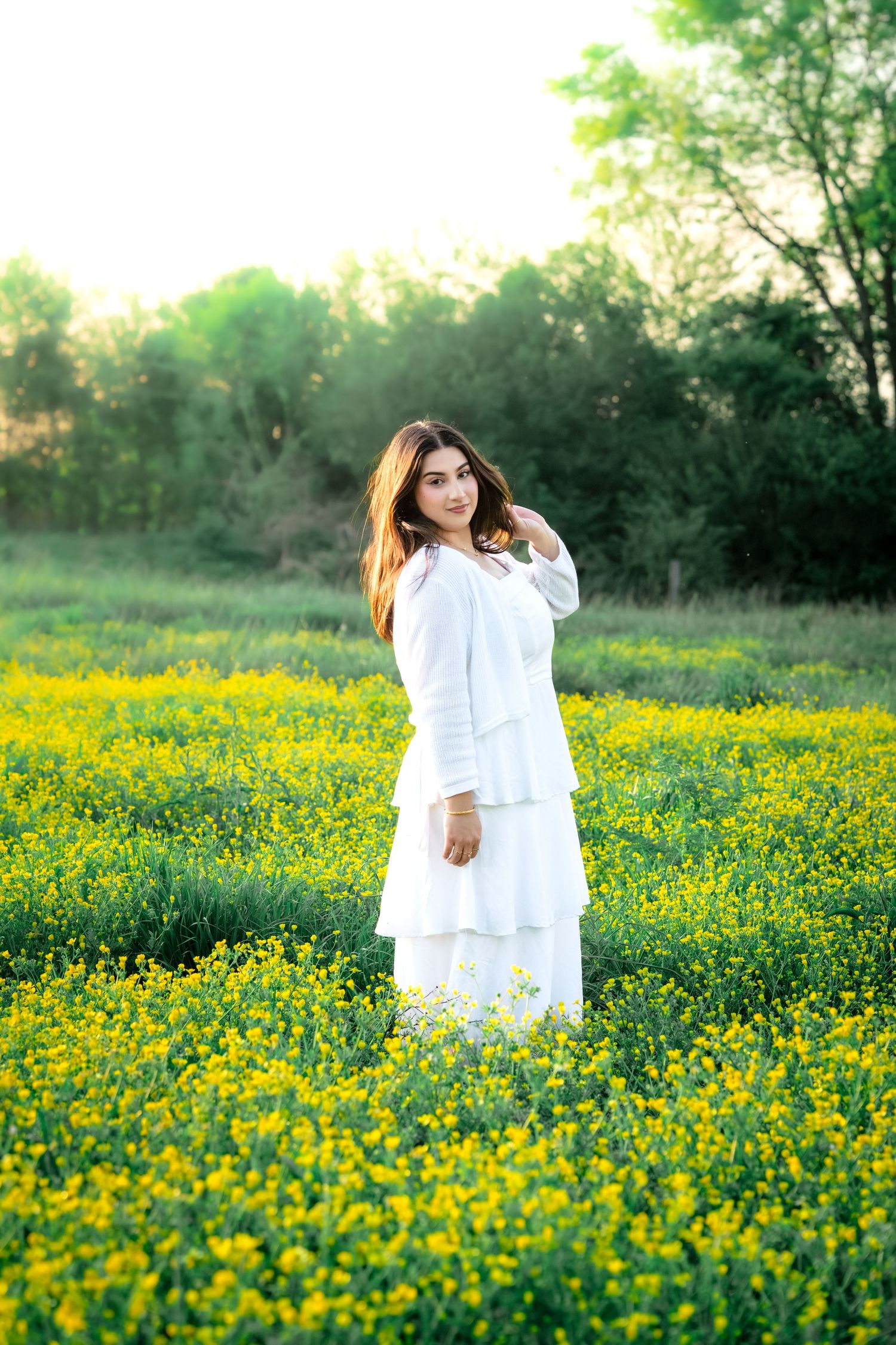 Young woman in white tiered dress standing in yellow wildflower field at golden hour.
