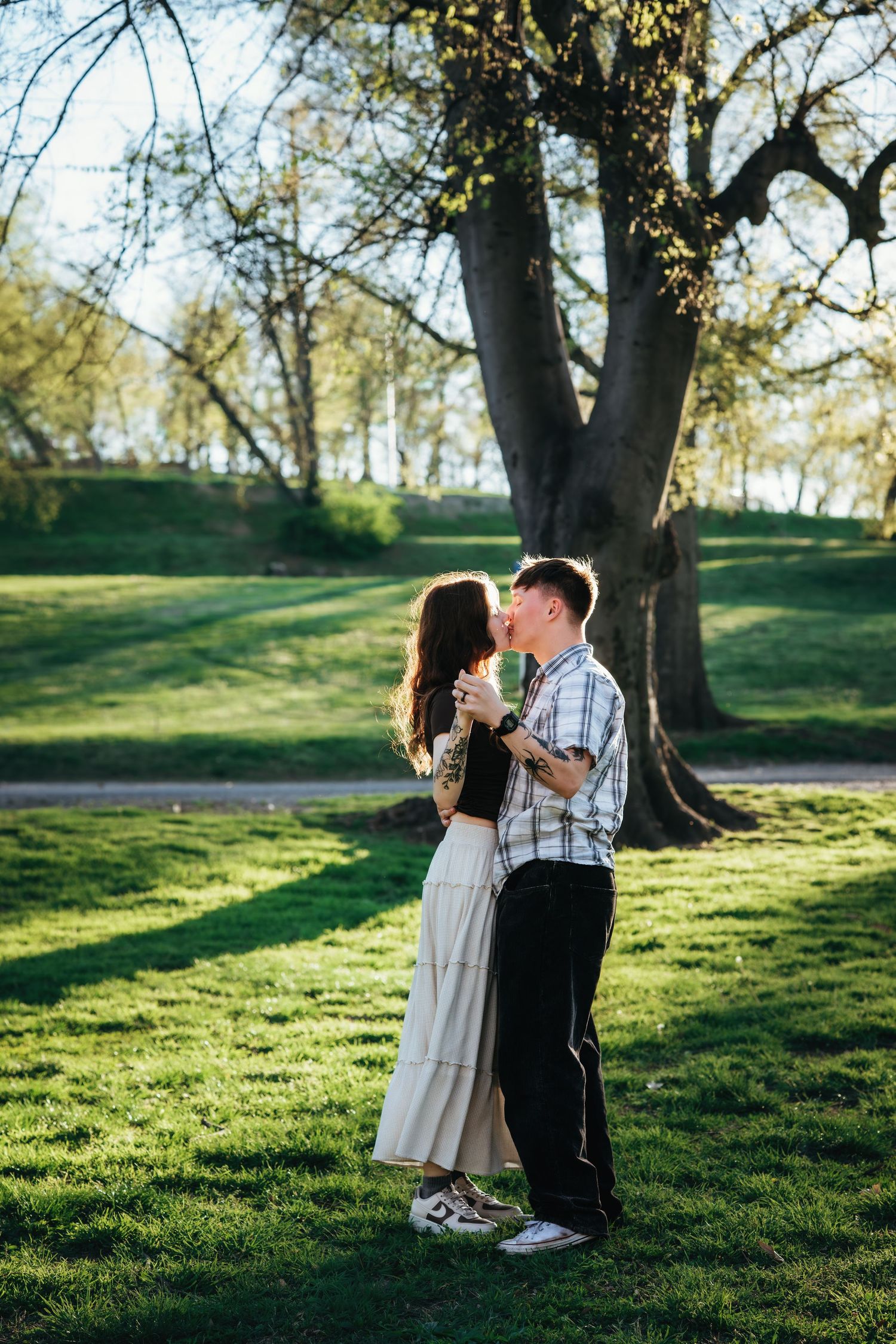 Couple sharing romantic kiss on lush green grass beneath large tree in sunny park.