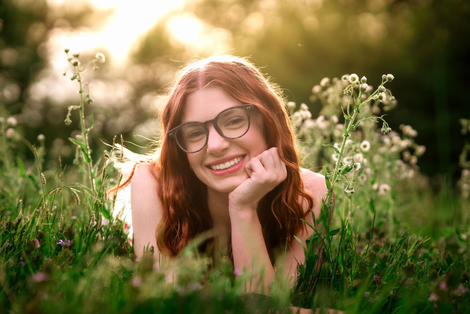 Smiling redhead woman with glasses lying in green grass field with wildflowers in warm sunlight.