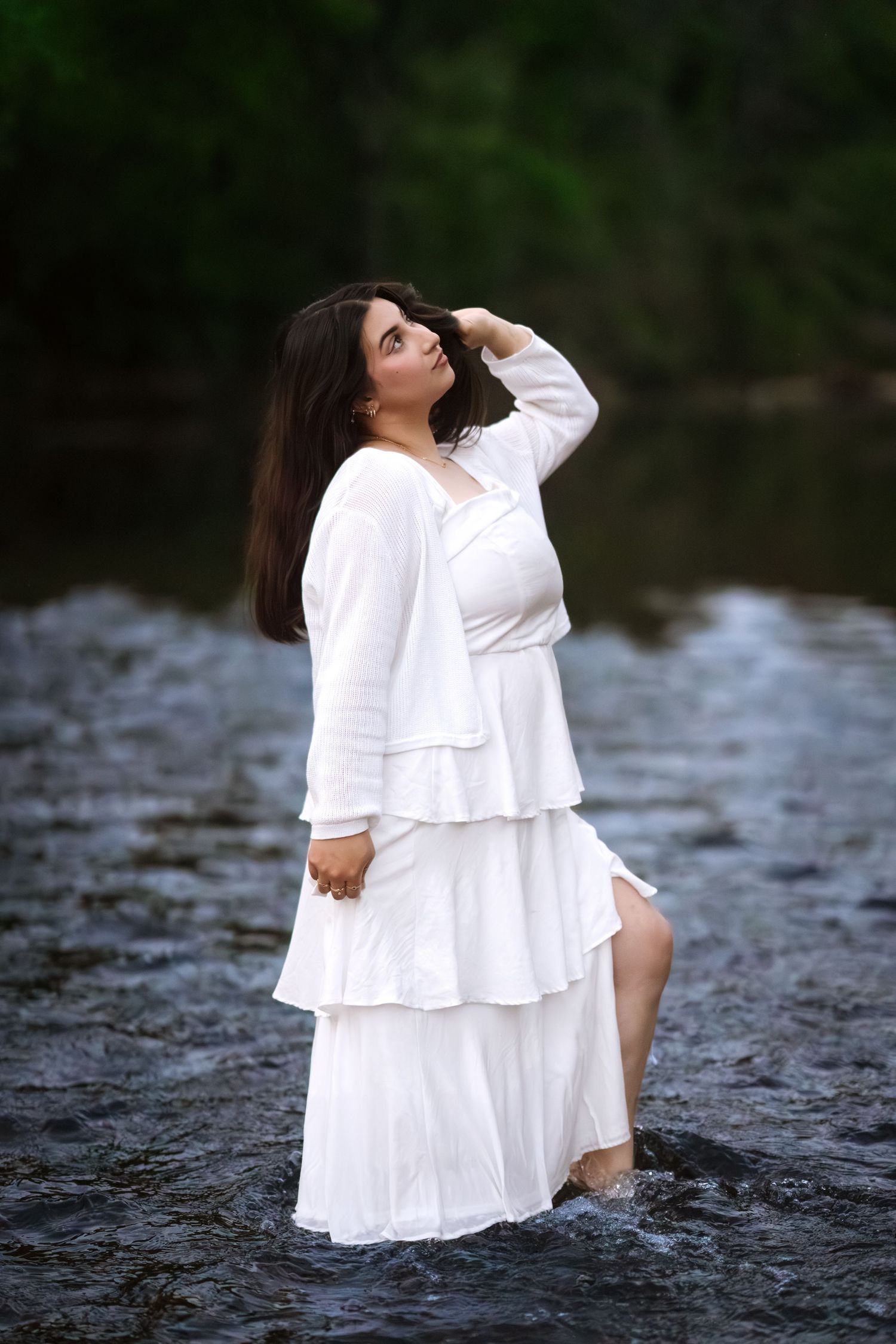 Woman in flowing white tiered dress standing in a serene river, hand in hair, surrounded by lush greenery.