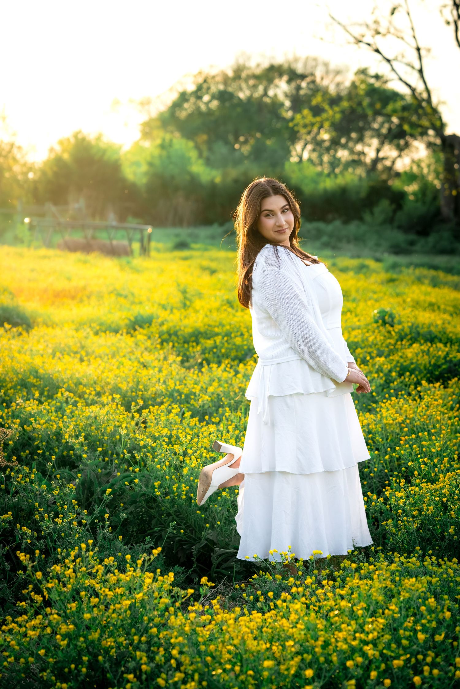 Young woman in white tiered dress holding heels in a sunny yellow wildflower field at golden hour.