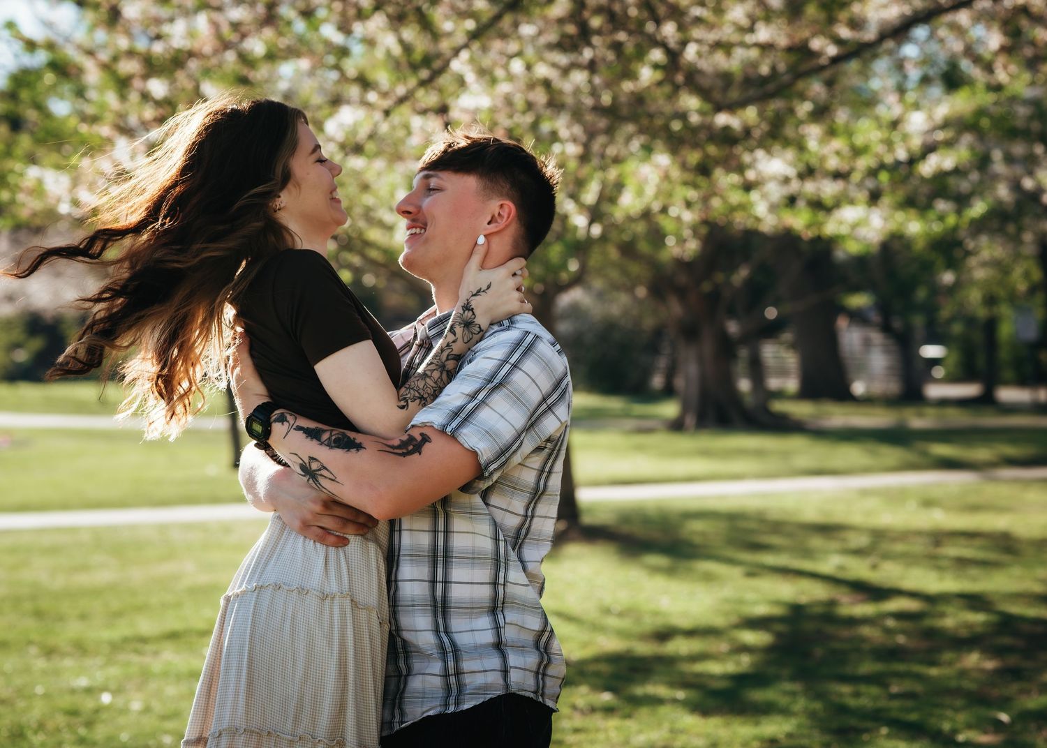 Couple embracing and laughing in a sunny park with blooming cherry blossom trees in the background.