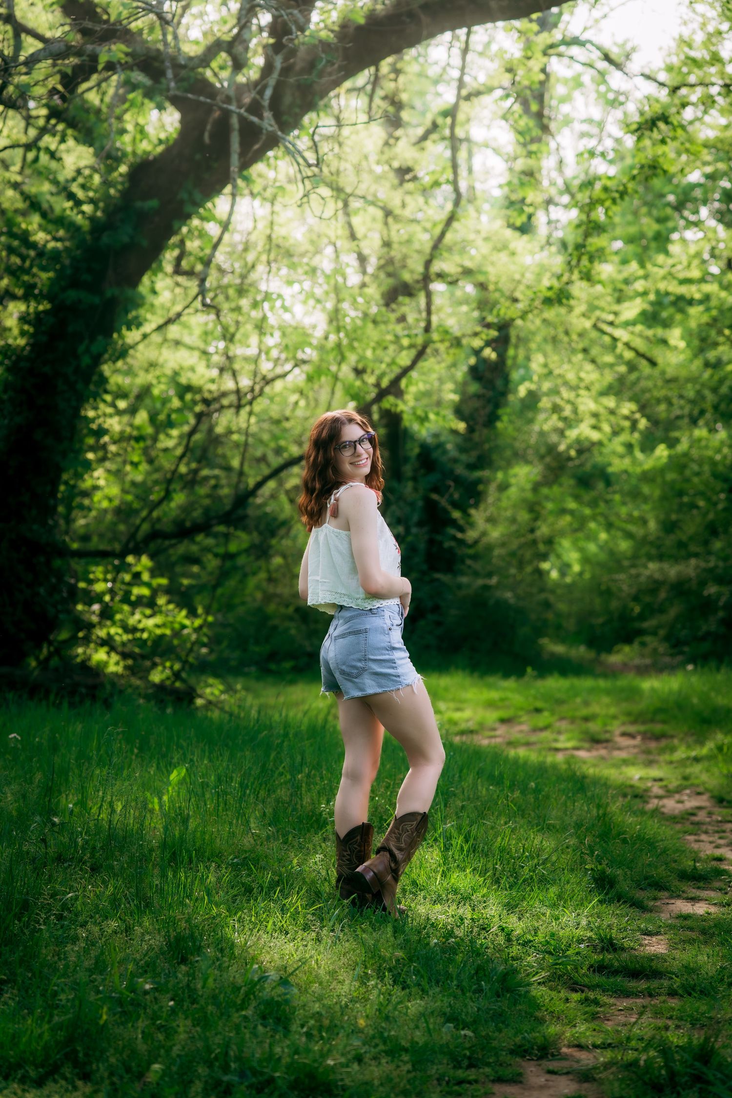 Young redhead woman in denim shorts and cowboy boots posing in a lush green forest clearing.