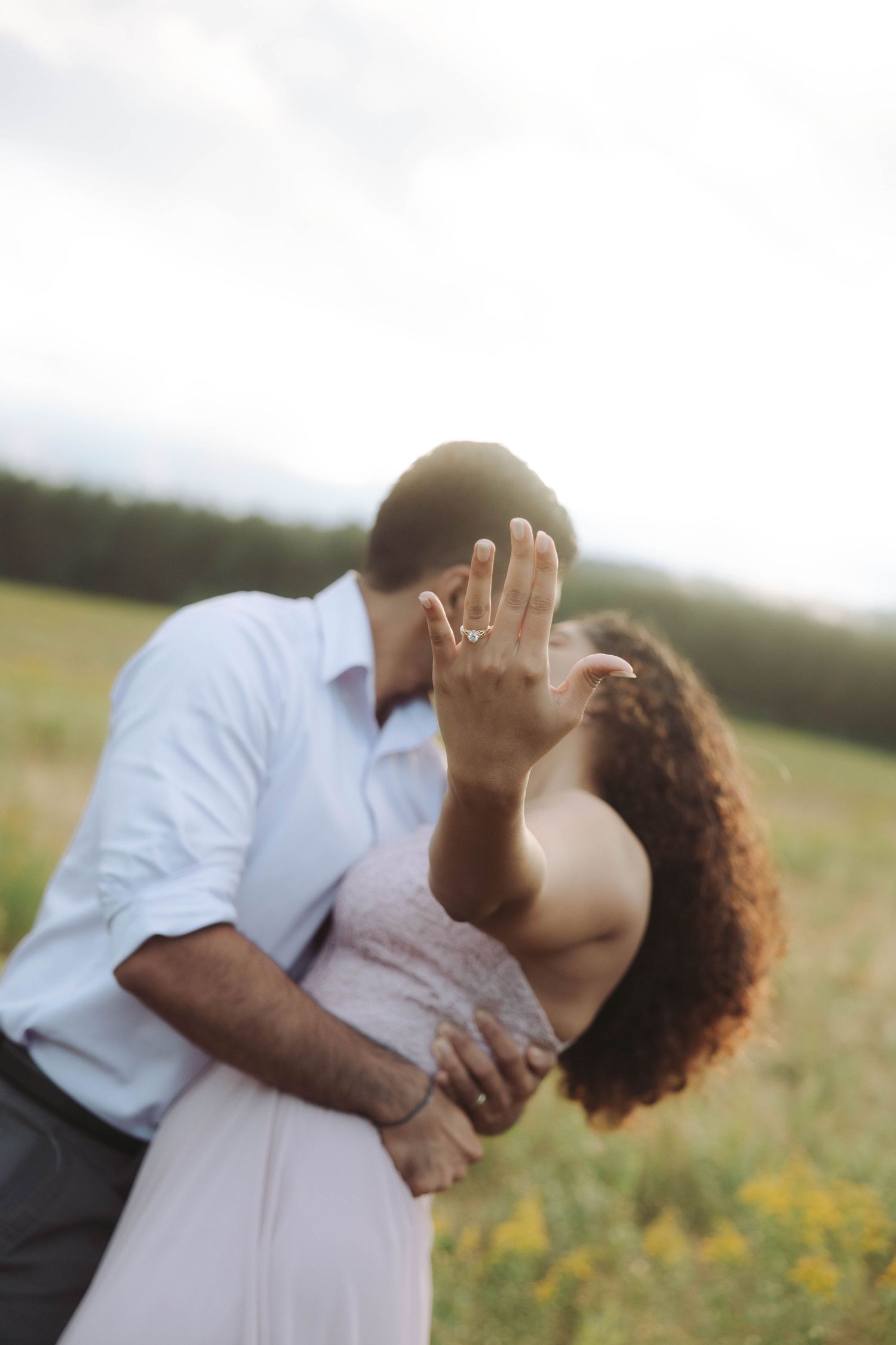 Engaged couple kissing in field while woman shows off diamond engagement ring to camera.