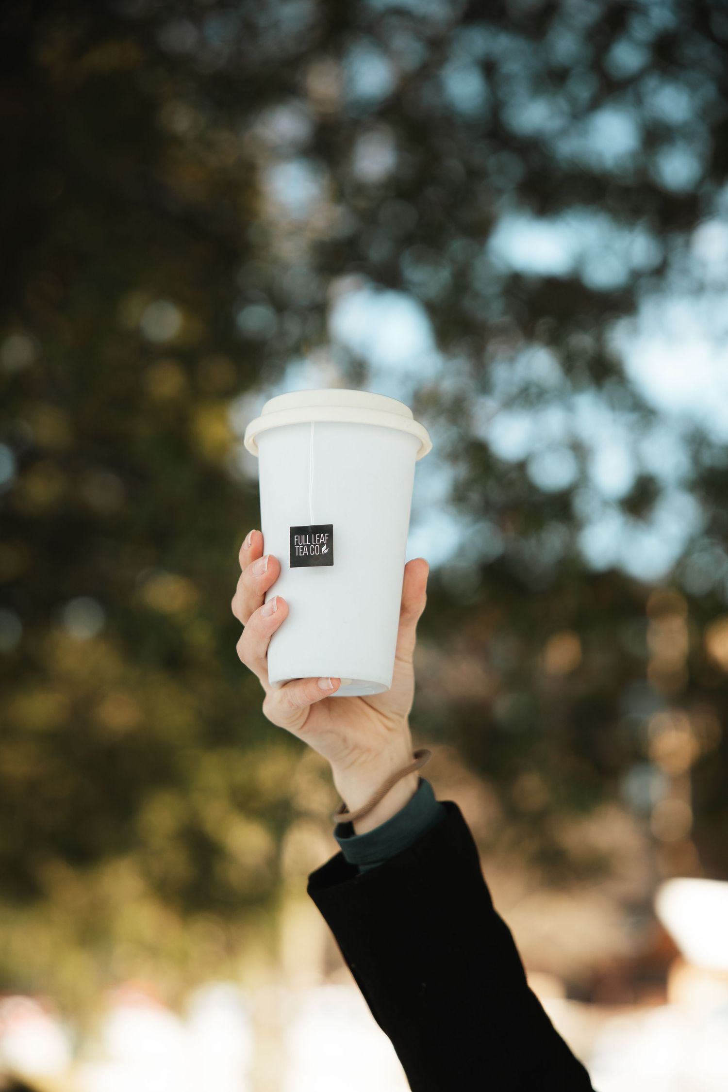Hand raised holding a white reusable cup with a small label against a blurred outdoor background.