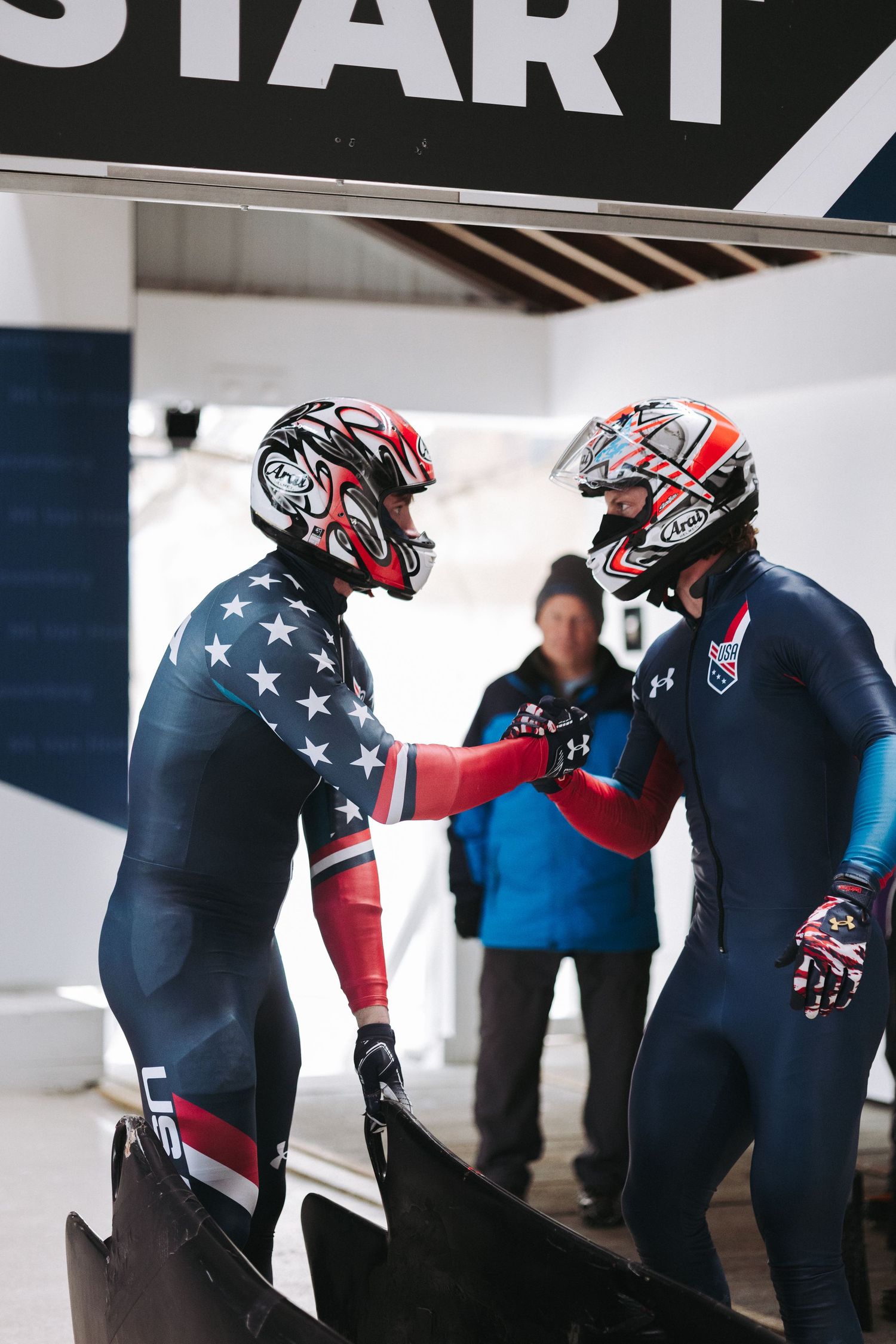 Two USA bobsled athletes in star-spangled suits fist bumping at competition start line.