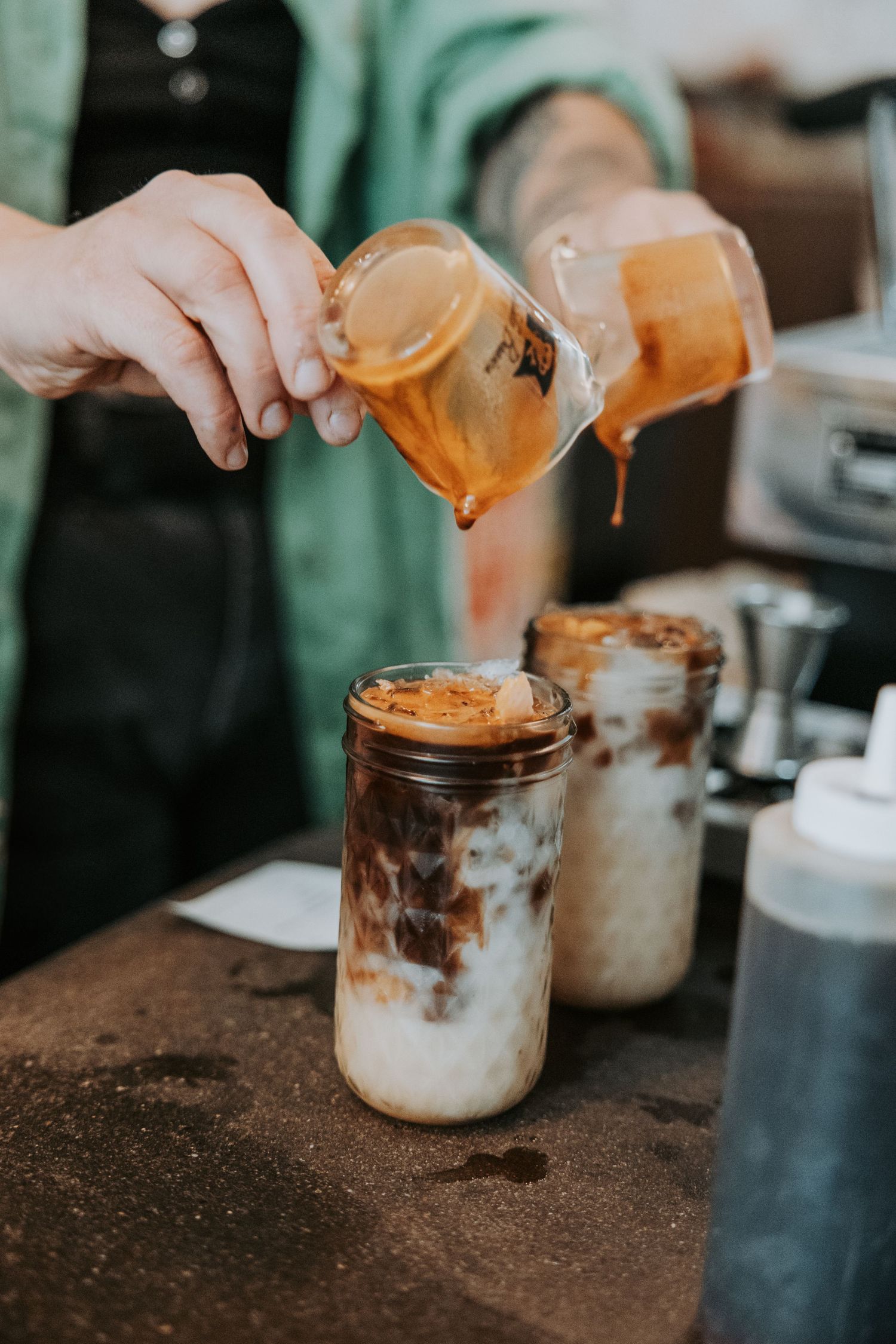 Barista pouring espresso shot into an iced latte in a mason jar on a coffee shop counter.