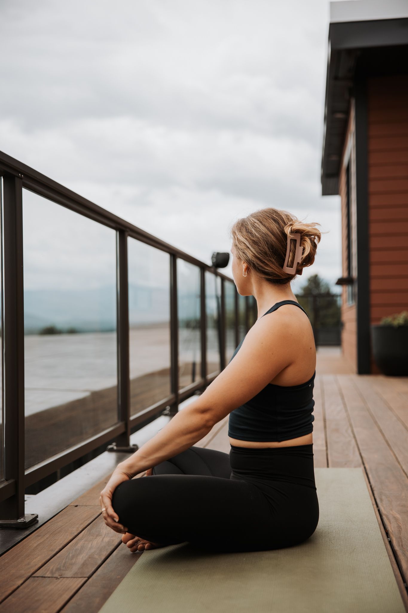 Blonde woman meditating in lotus pose on yoga mat on wooden deck overlooking mountains.