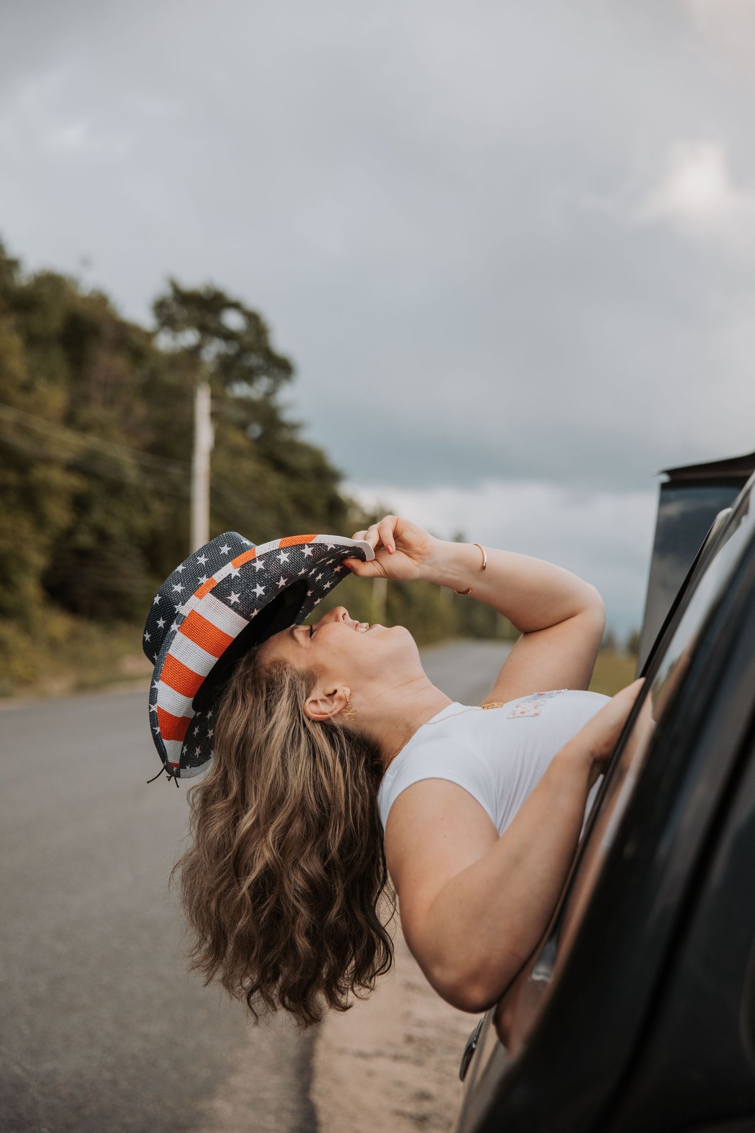 Young woman leaning out of a car window wearing an American flag bucket hat on a road.
