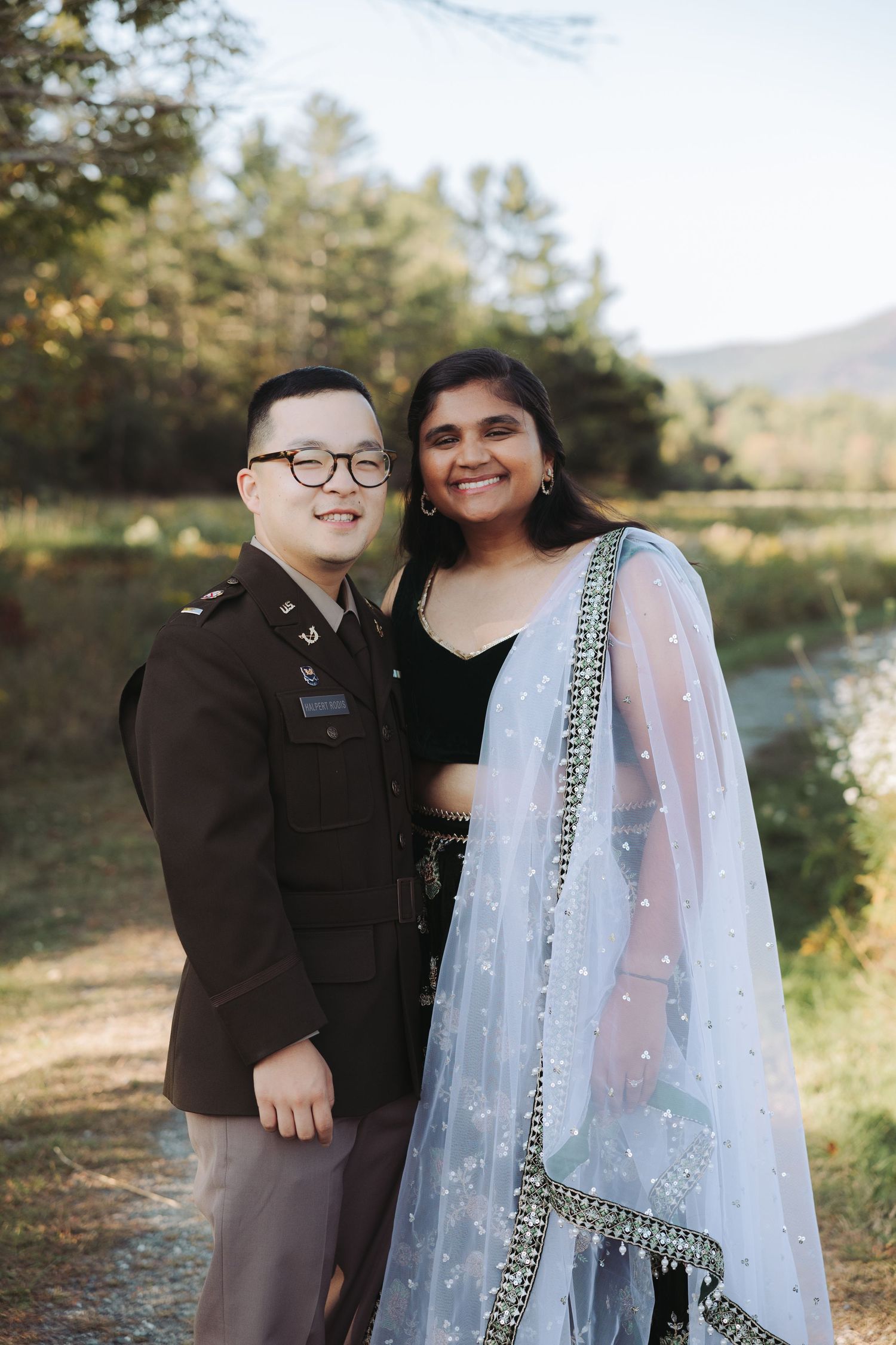 Smiling couple posing outdoors, man in military uniform and woman in elegant blue Indian lehenga.
