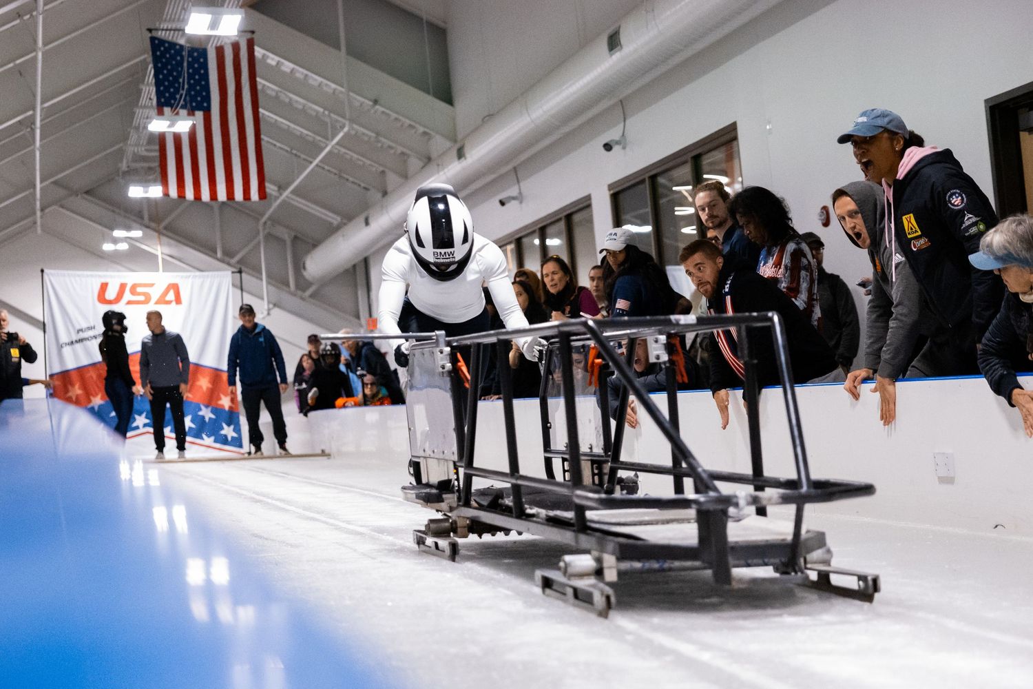 USA bobsled athlete in aerodynamic helmet pushes off at indoor training facility with coaches and American flag in background.