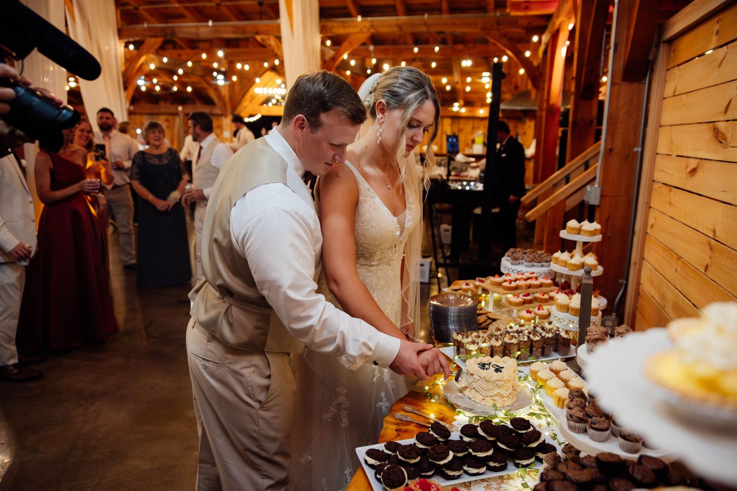 A bride and groom cut their wedding cake at a rustic barn reception with dessert table.