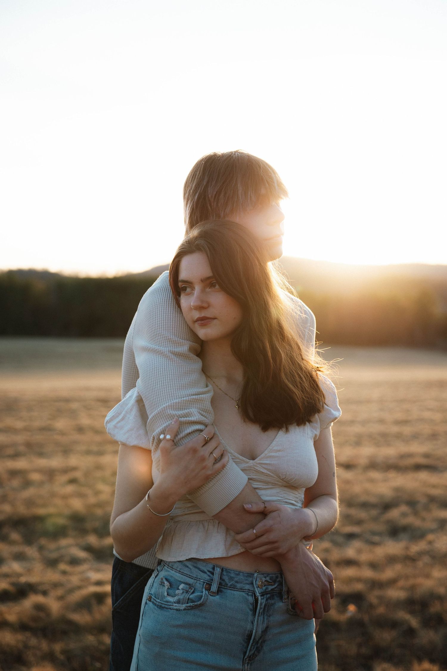 Man embracing woman from behind in a golden sunset field, both wearing casual white tops.