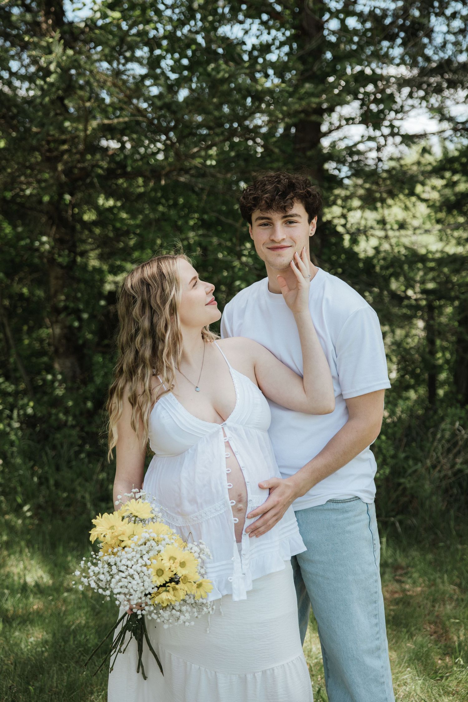 Pregnant woman in white dress holding yellow flowers stands with young man in white shirt outdoors.