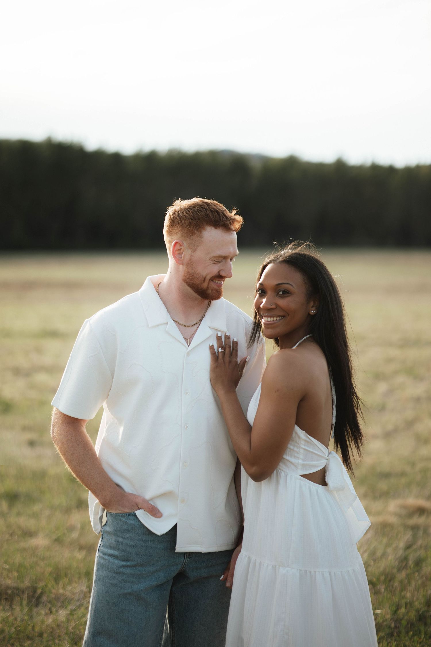 Interracial couple smiling together in a sunlit field, woman in white sundress during golden hour.