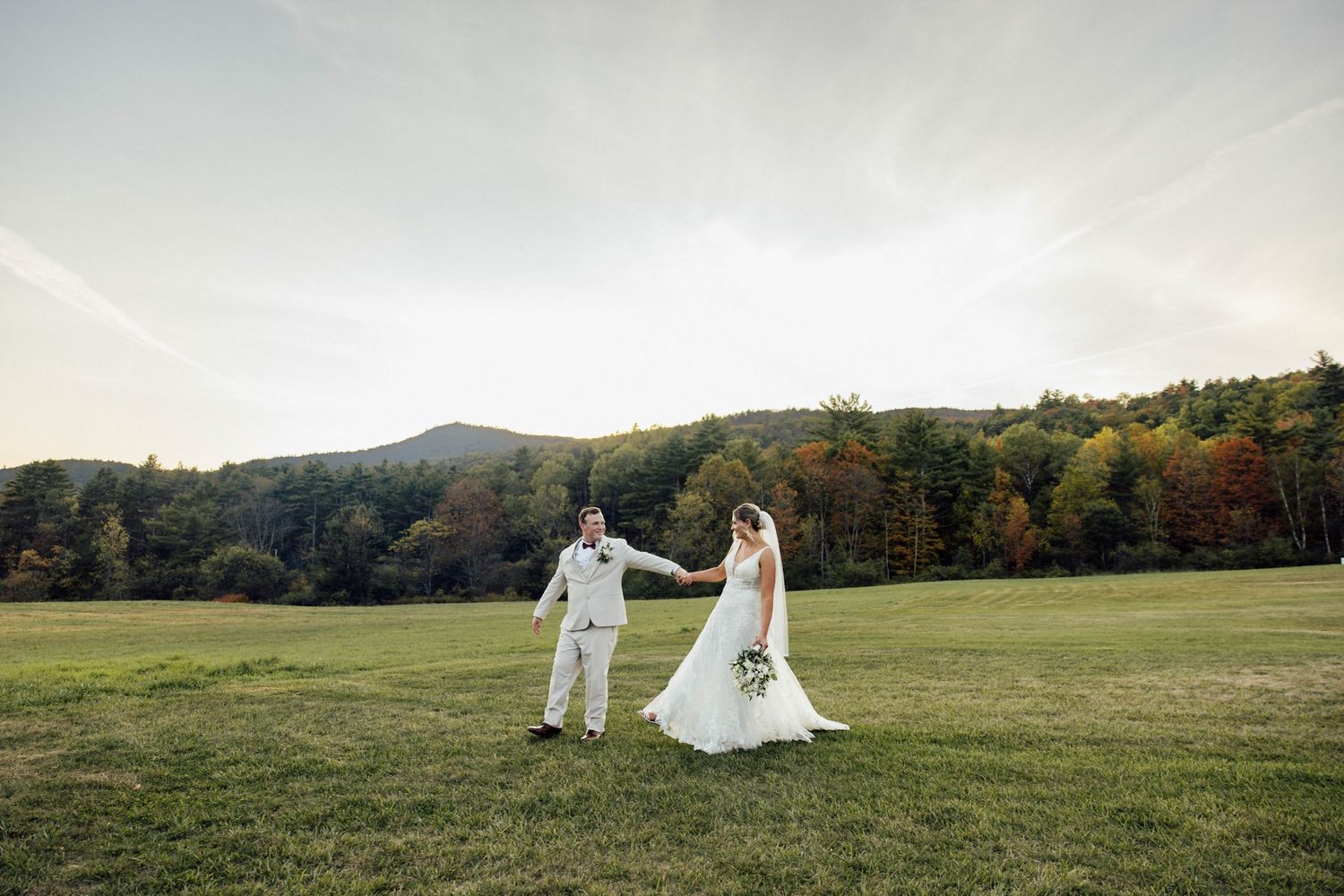 Bride and groom holding hands walking through green meadow with colorful autumn mountain forest backdrop at outdoor wedding.