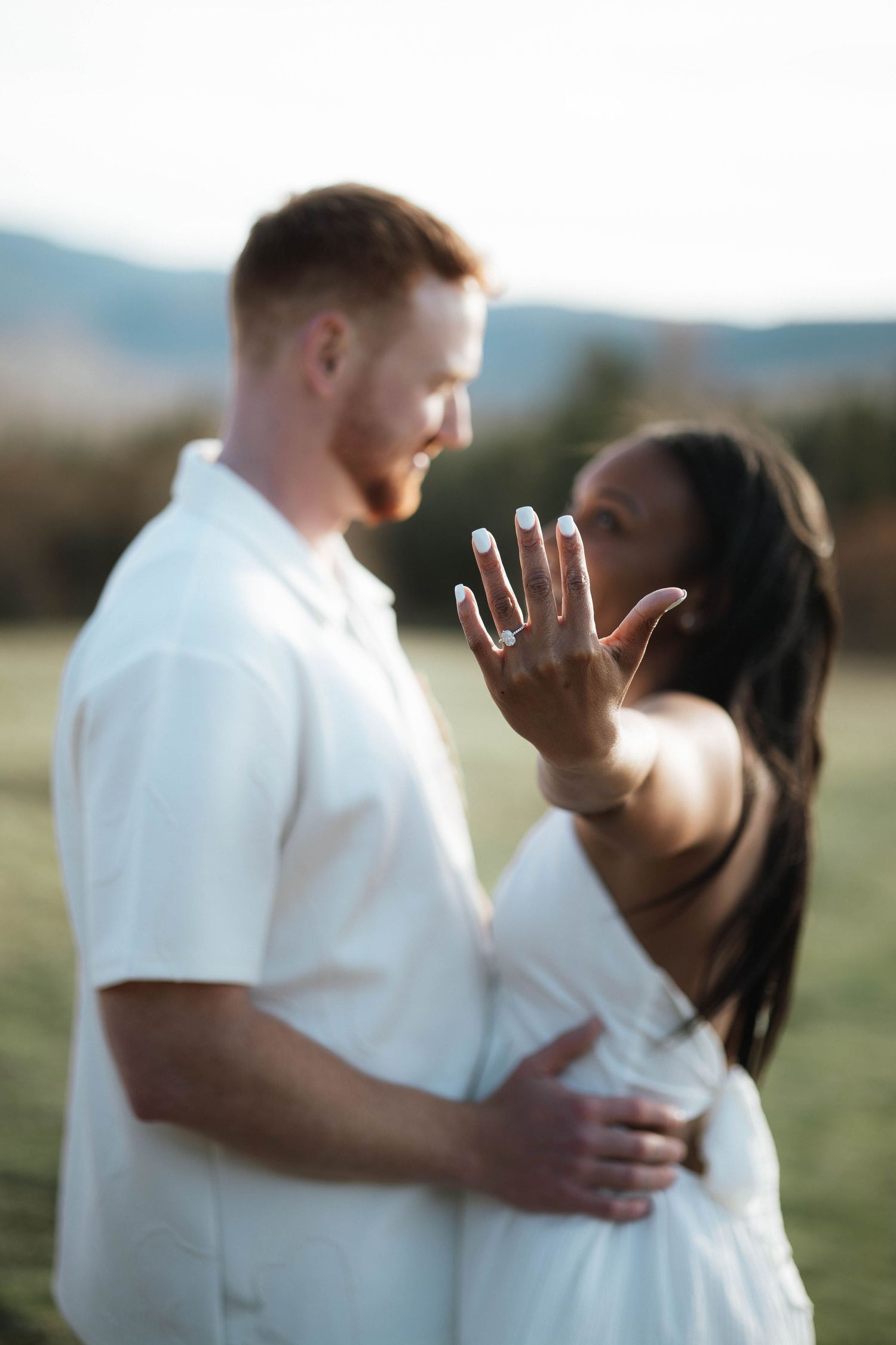 Interracial couple in white outfits showing engagement ring outdoors at golden hour.