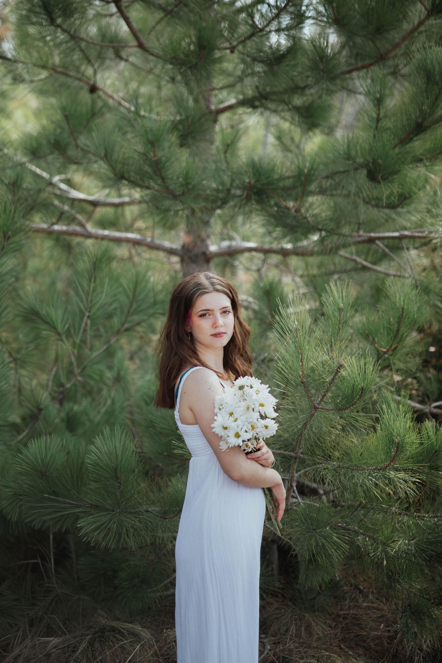Young woman in white dress holding white flowers standing among green pine trees outdoors.