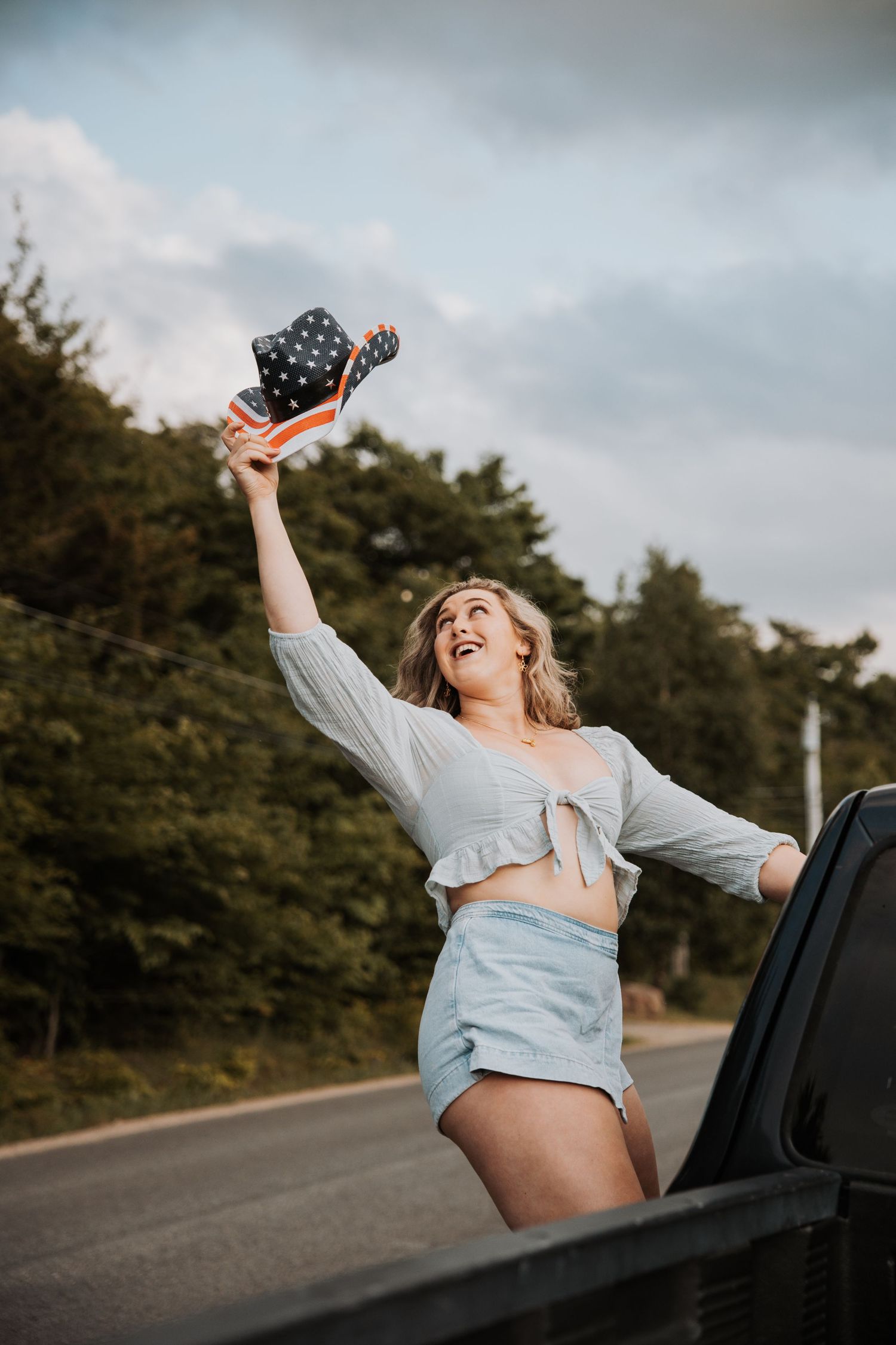Joyful woman holding an American flag hat out of a car door on a tree-lined road.