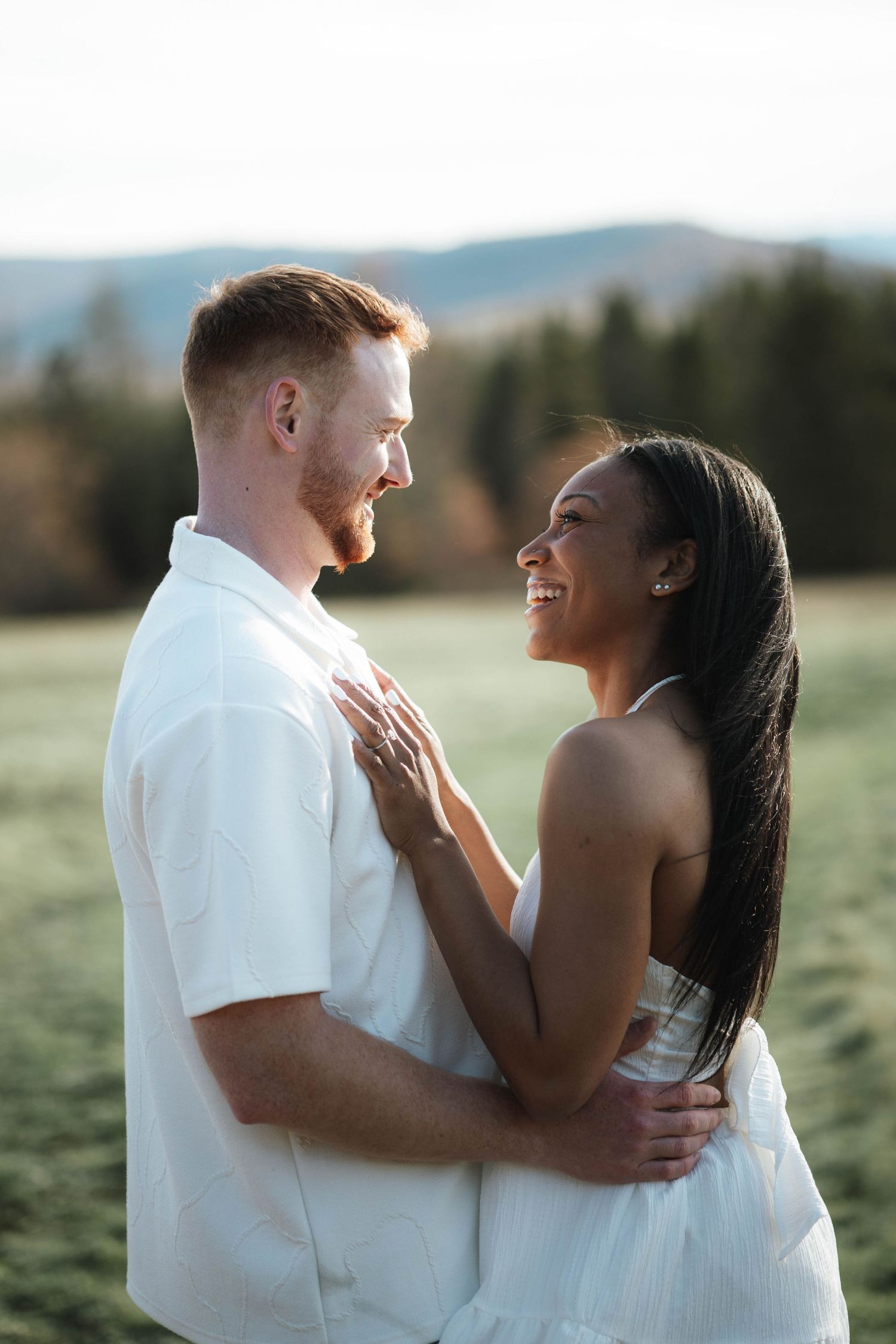 Interracial couple embracing and smiling at each other in a green field, both wearing white outfits with mountains in background.