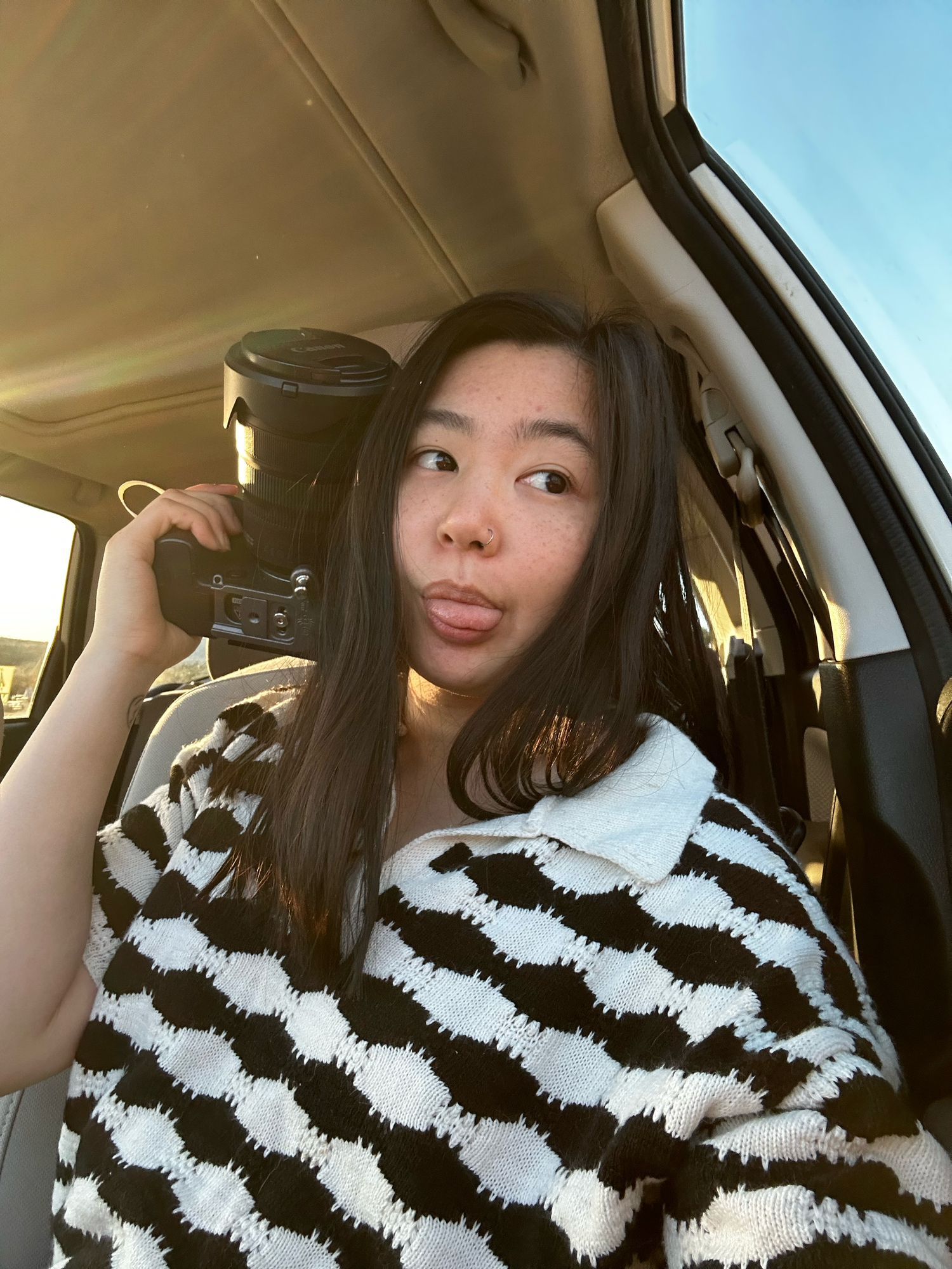 Young woman in black and white patterned sweater holding a camera in a car at golden hour.