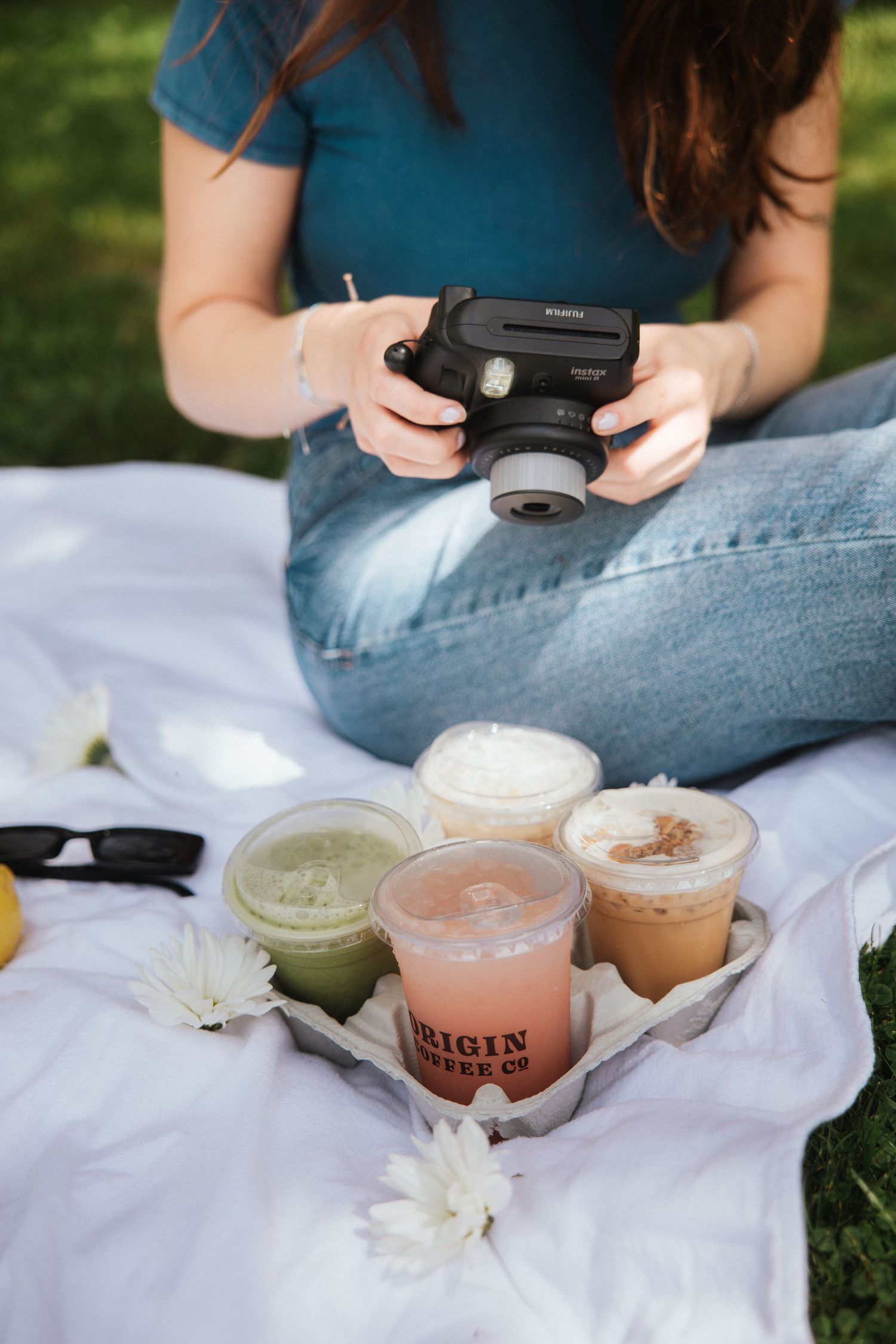 Woman photographing Origin Coffee Co drinks on a white blanket outdoors with an Instax camera.