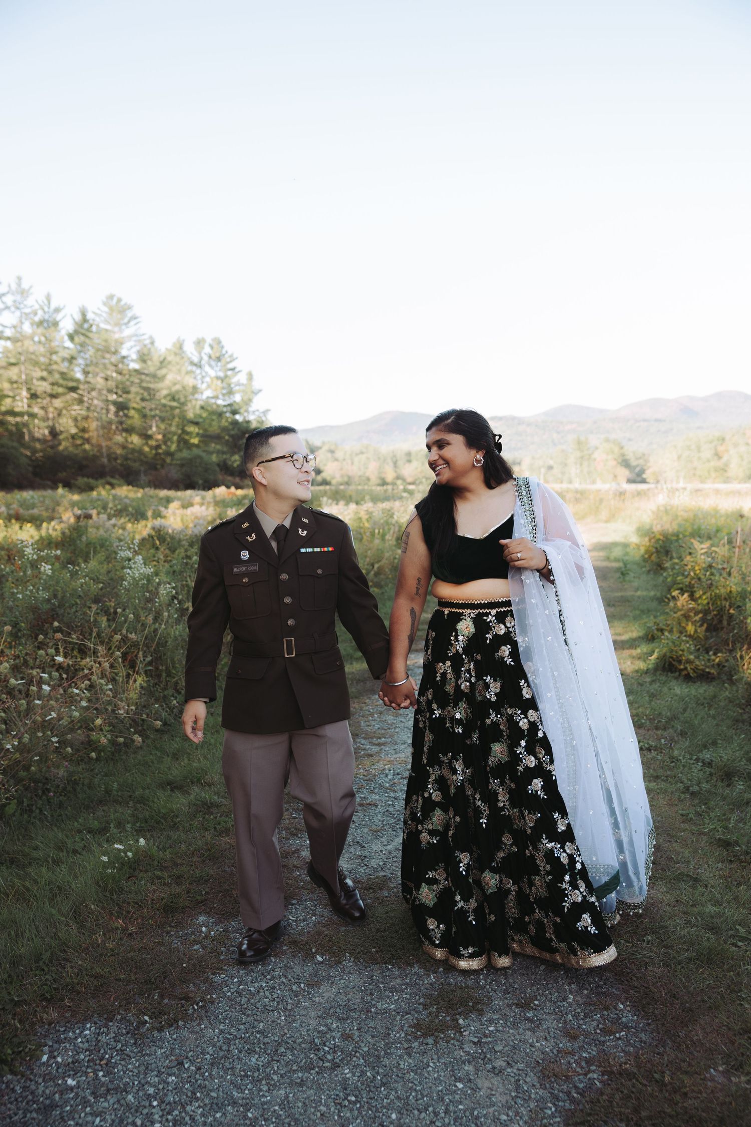 Couple walking hand-in-hand outdoors, man in military uniform and woman in black floral lehenga with blue dupatta.