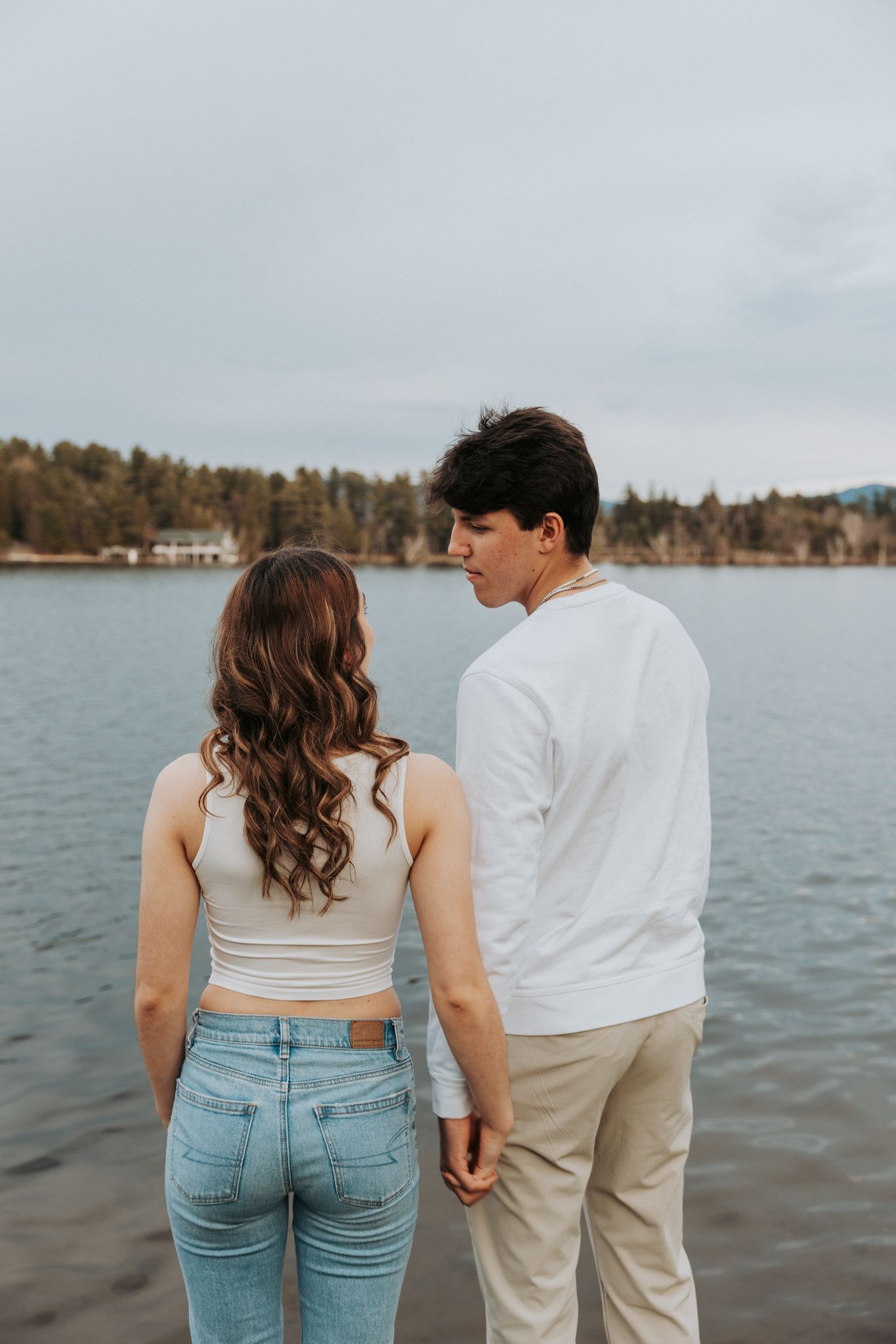 Couple holding hands facing a serene lake surrounded by autumn trees on a cloudy day.