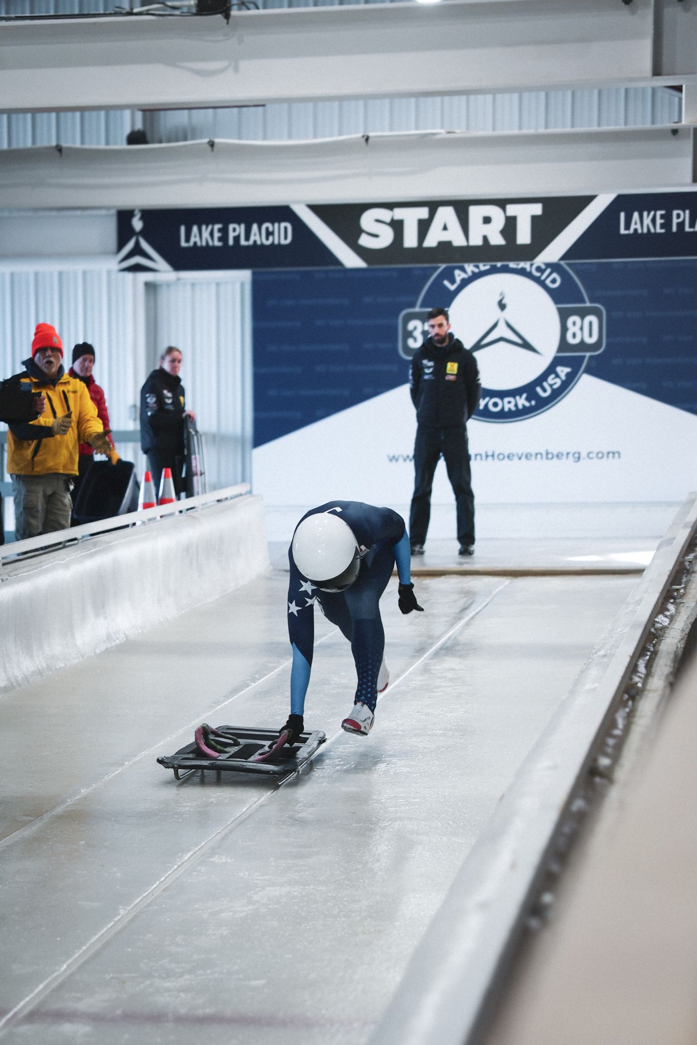 Athlete pushes skeleton sled at Lake Placid START line in New York, USA, with coaches watching at indoor ice track.