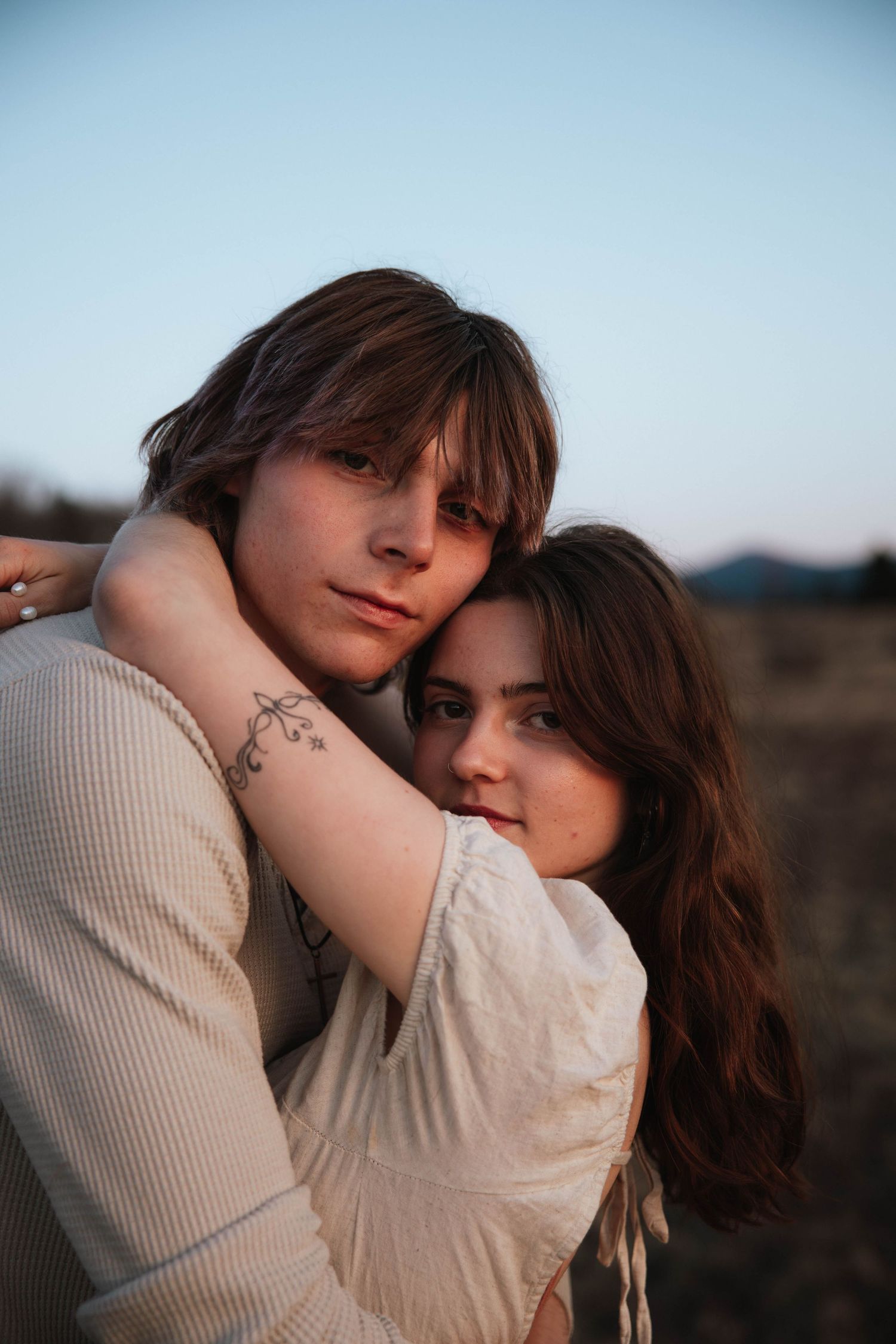 Young couple embracing outdoors at dusk, woman with floral arm tattoo hugging man in soft golden light.