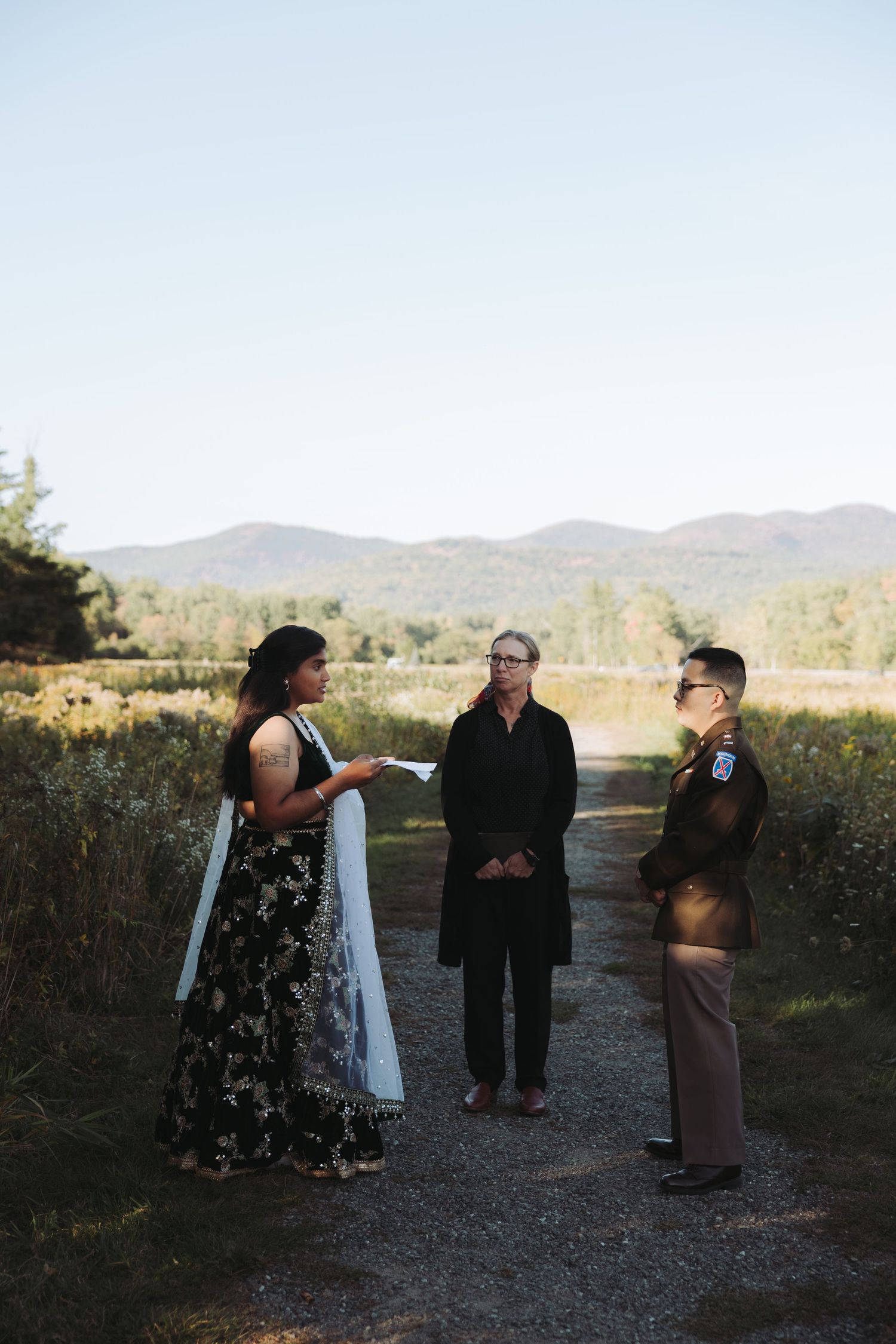 Three people stand on a gravel path during an outdoor wedding ceremony with mountain views.