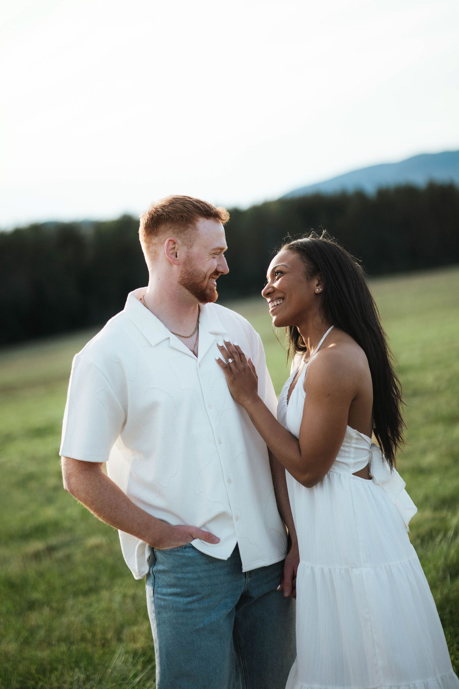 Interracial couple smiling at each other in a green field, wearing white outfits, with mountains in the background.