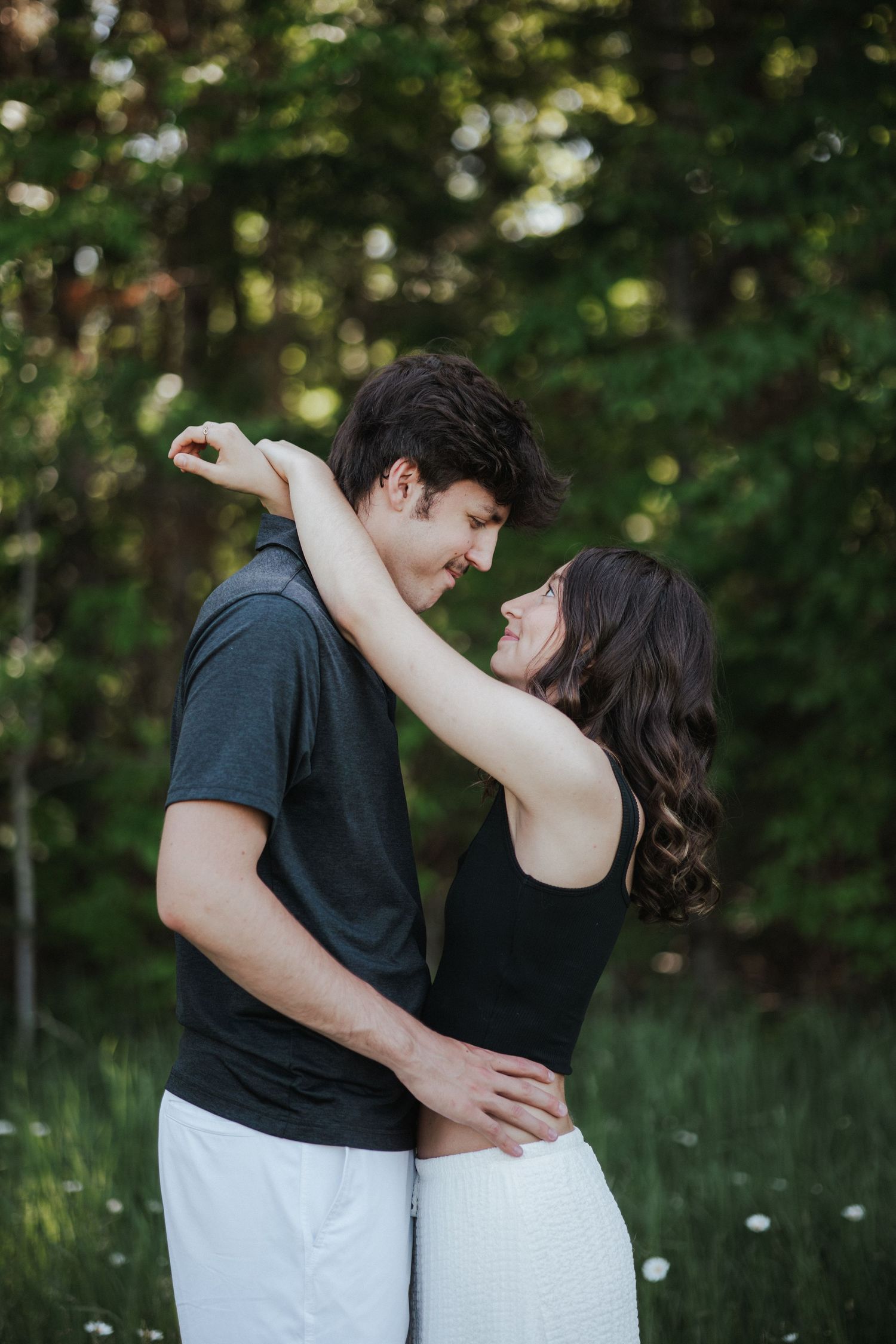 A couple embraces and gazes at each other lovingly outdoors, surrounded by lush green trees in a romantic setting.